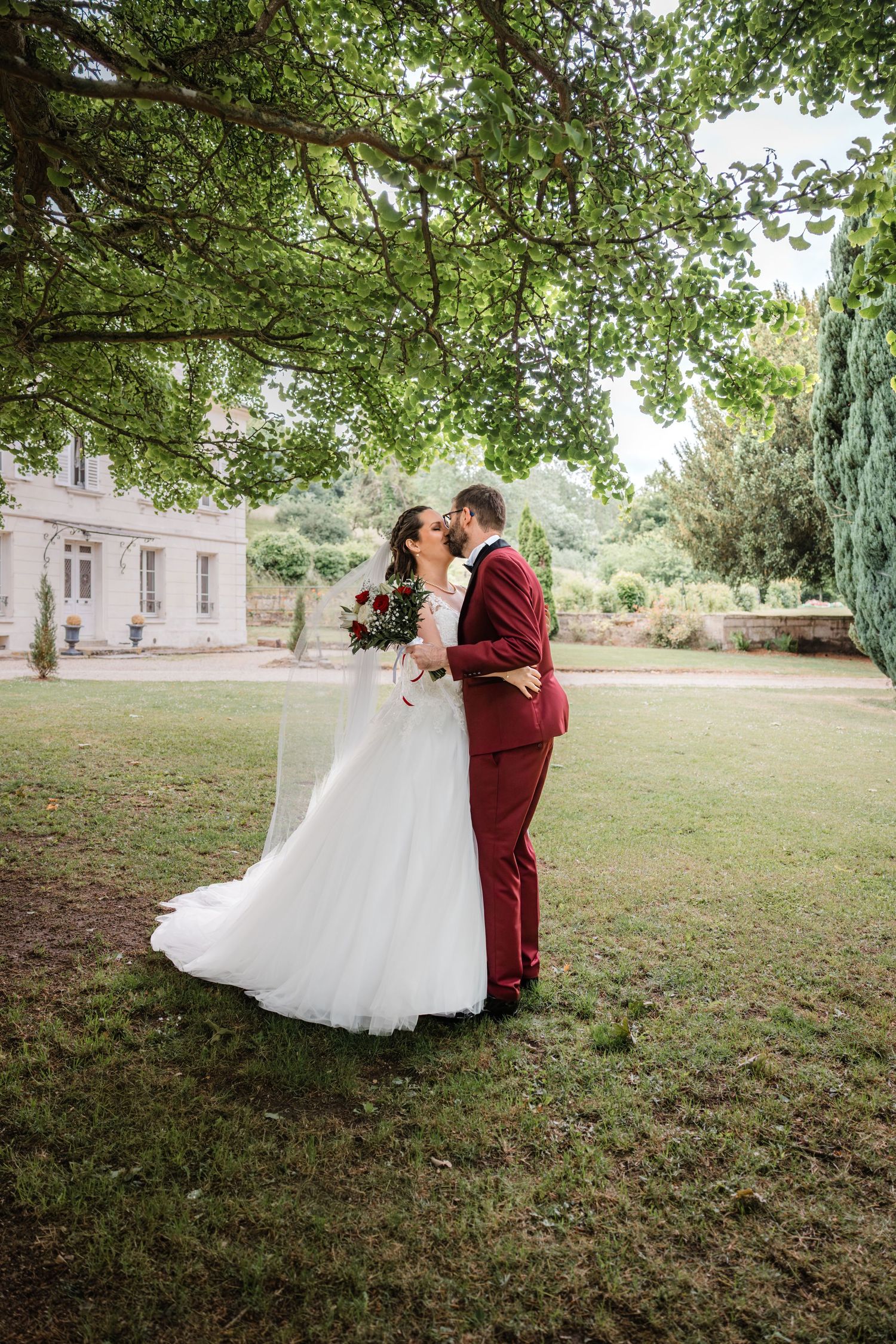 Couple de mariés romantiques partageant des moments tendres dans un jardin avec un élégant costume bordeaux et une robe blanche.