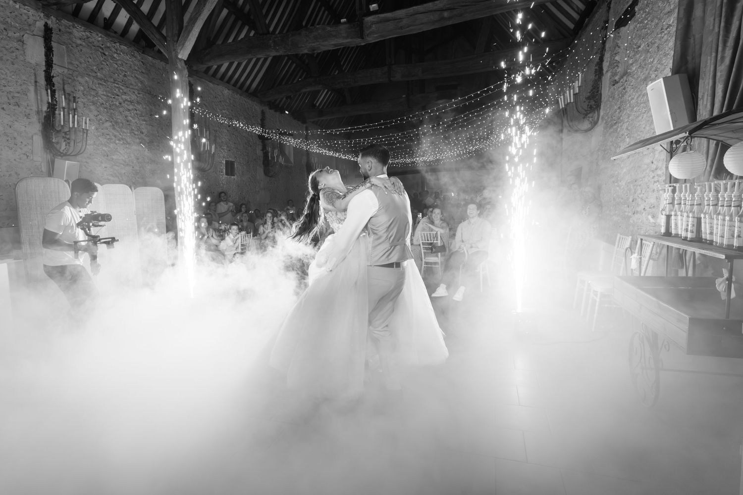 Photo en noir et blanc des mariés partageant leur première danse dans une grange rustique, avec guirlandes lumineuses et brume théâtrale.