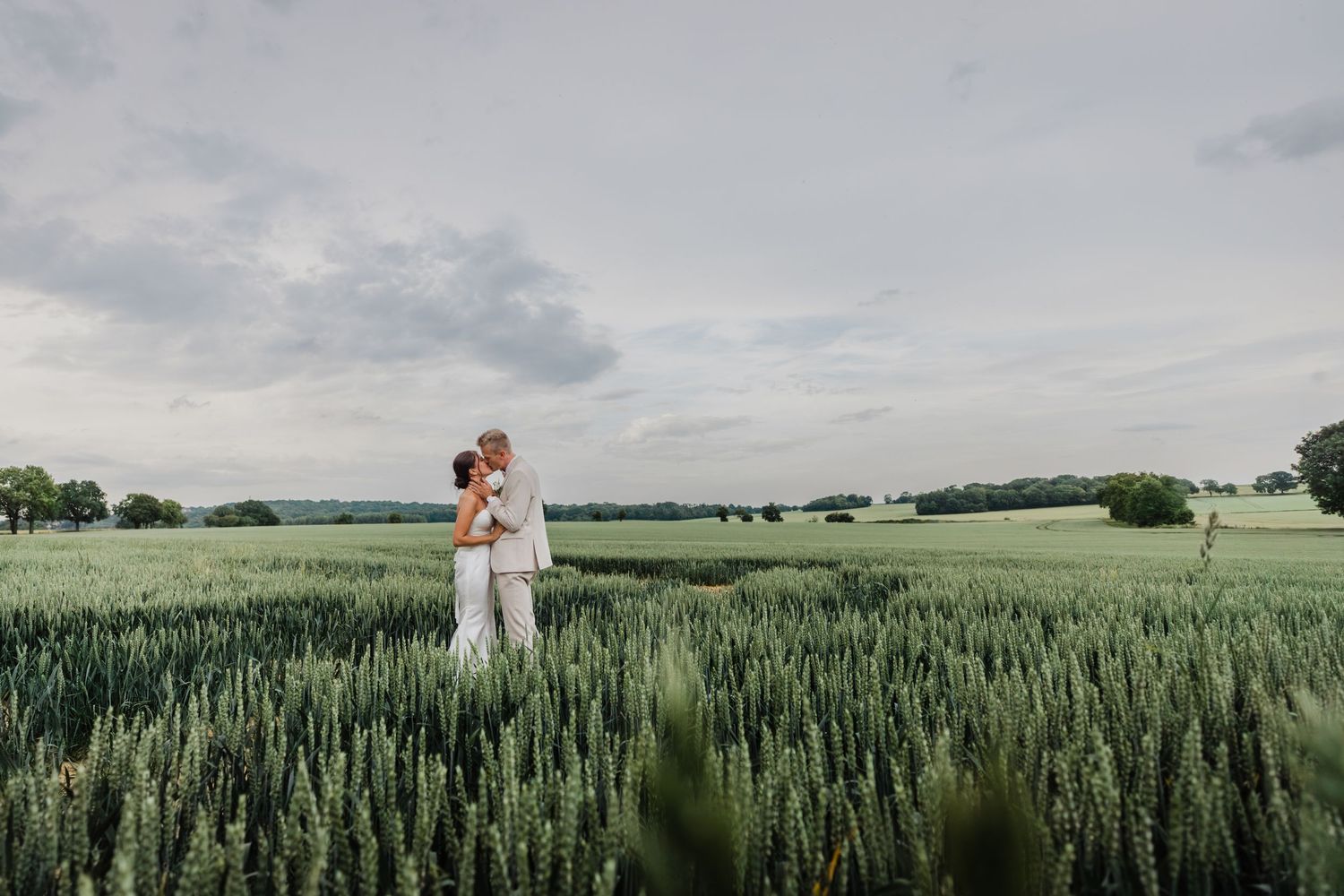 Un couple s'embrasse amoureux entouré d'un champ de blé vert luxuriant sous un ciel d'été nuageux.