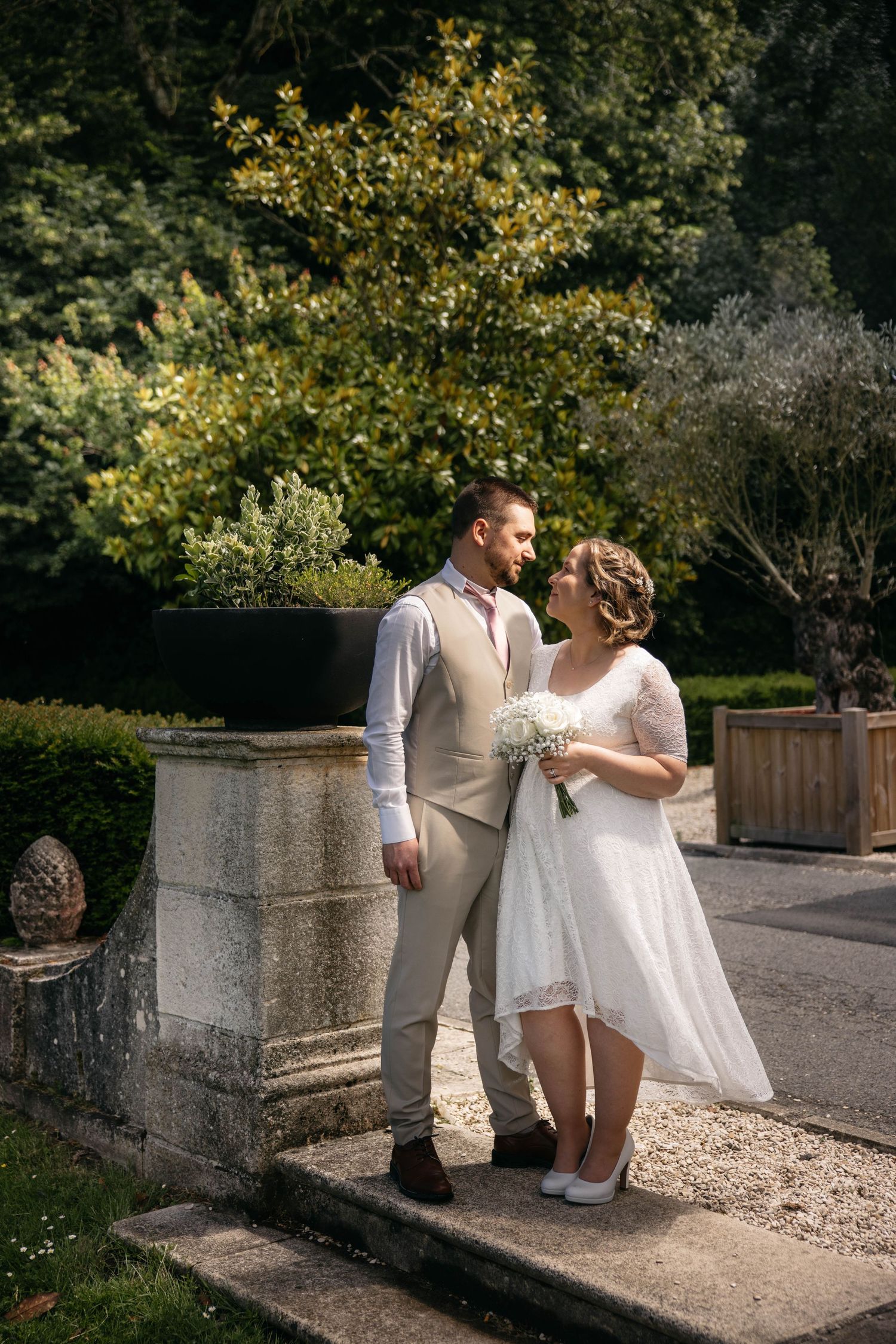 La mariée en robe blanche, tenant son bouquet, se tient aux côtés du marié en costume beige, à l'extérieur, dans un cadre de jardin