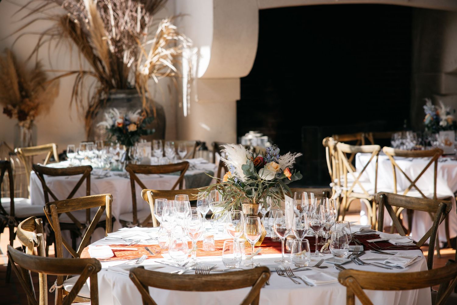 Tables de réception élégantes : chaises à dossier croisé, centres floraux, verrerie et pampa près d'une cheminée.