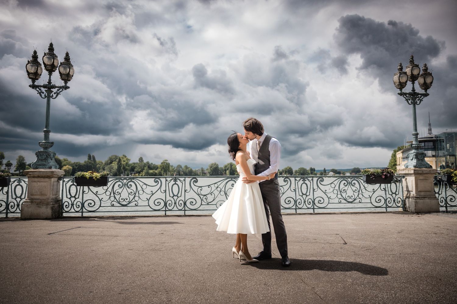 Un couple romantique s'embrasse sur un pont orné avec des balustrades en fer forgé sous un ciel orageux spectaculaire.