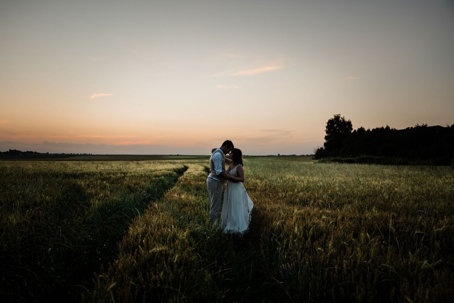 A couple embraces in a golden wheat field at sunset with silhouettes against the dusky sky.