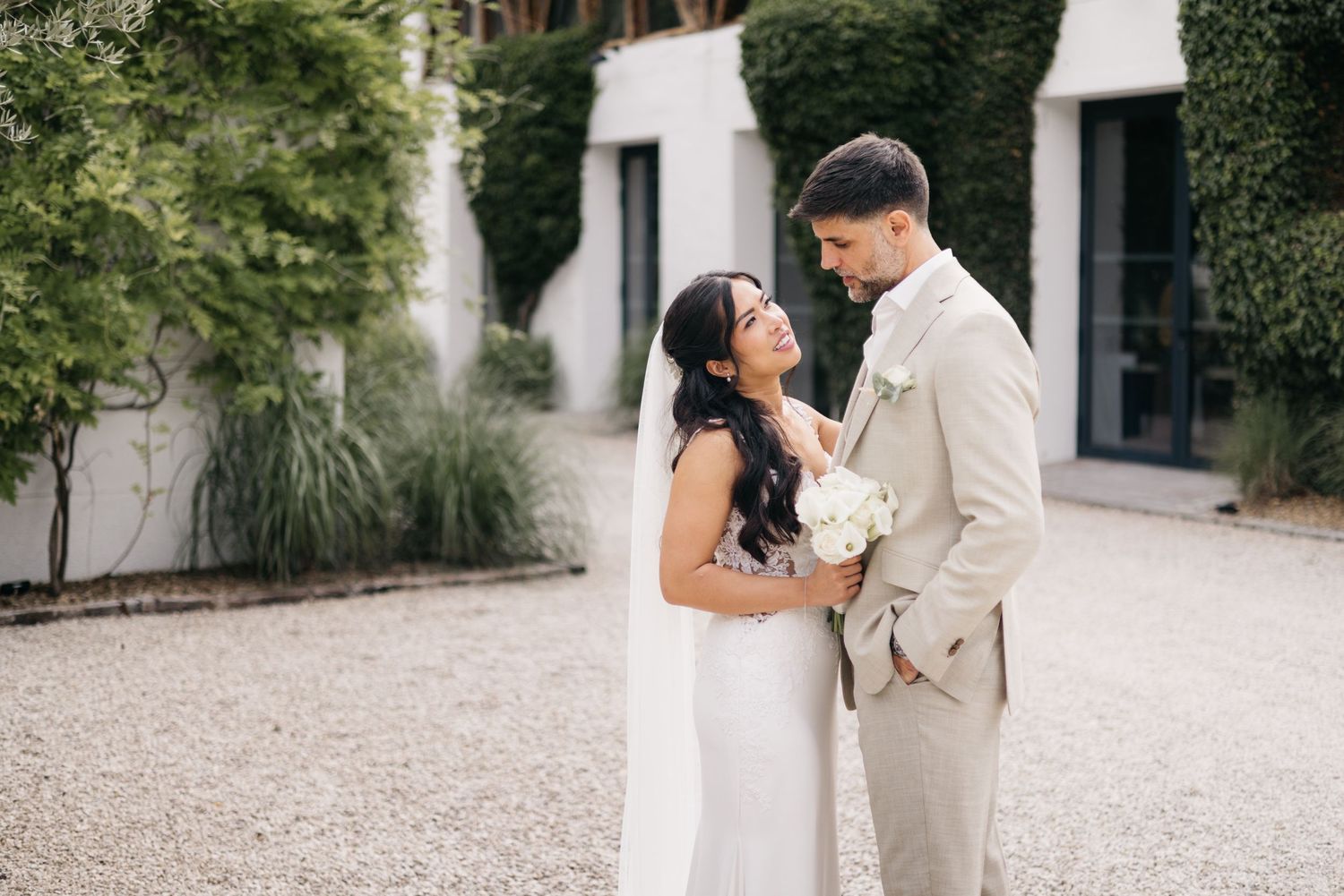 Les mariés partagent un tendre moment en plein air, elle vêtue d'une robe blanche et lui d'un costume beige, entourés d'une végétation
