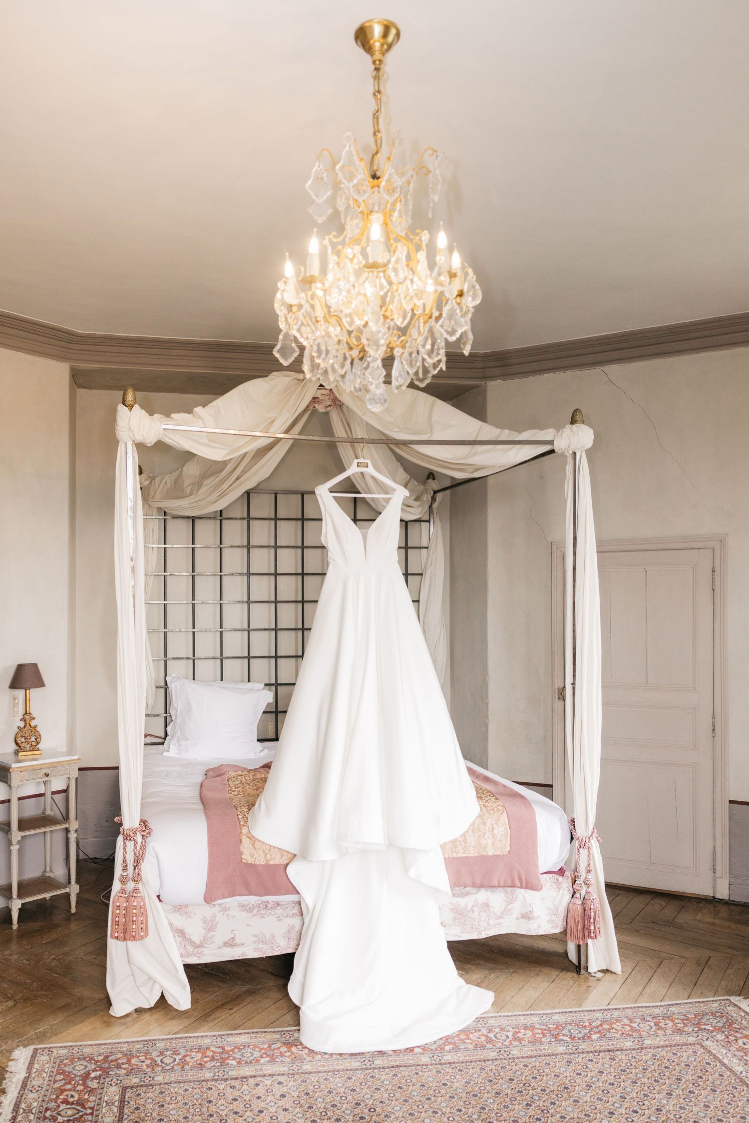 Robe de mariée blanche suspendue à un lit à baldaquin sous un lustre en cristal, dans une chambre élégante d’un château français.