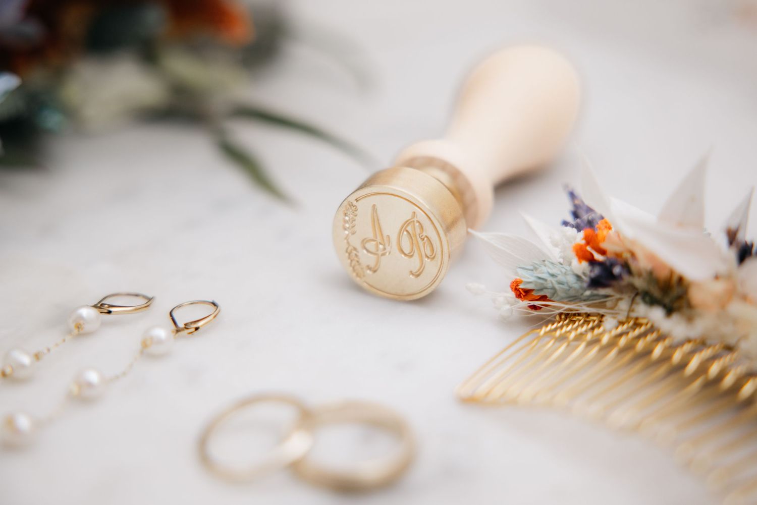 Mise à plat de mariage : cachet de cire, alliances dorées, boucles en perles et peigne à fleurs sur marbre.