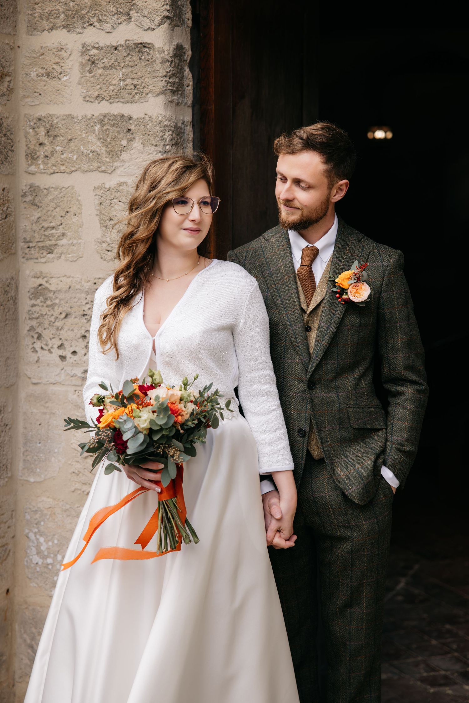 Un couple en tenue de mariage pose contre un mur de pierre avec une mariée tenant un bouquet de couleur automne avec un ruban orange.