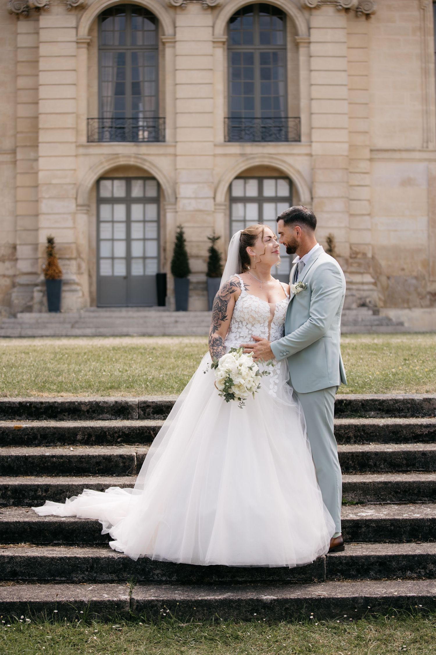 Les mariés s'enlacent sur des marches en pierre devant un grand bâtiment, elle tient un bouquet blanc dans une élégante robe de bal.