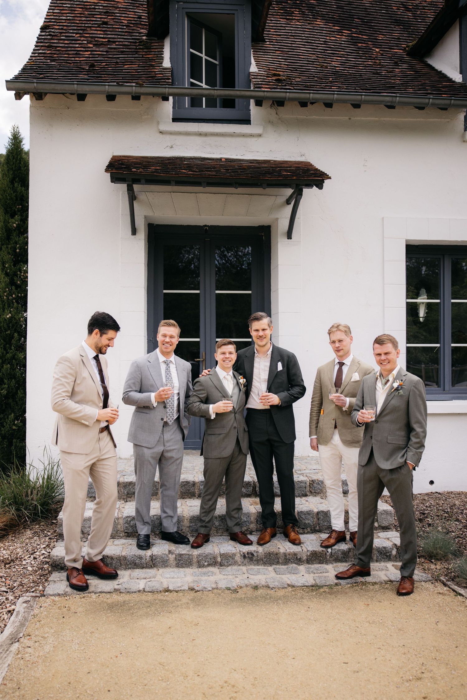 Six garçons d'honneur en costume posent sur des marches en pierre devant un lieu de style français blanc.