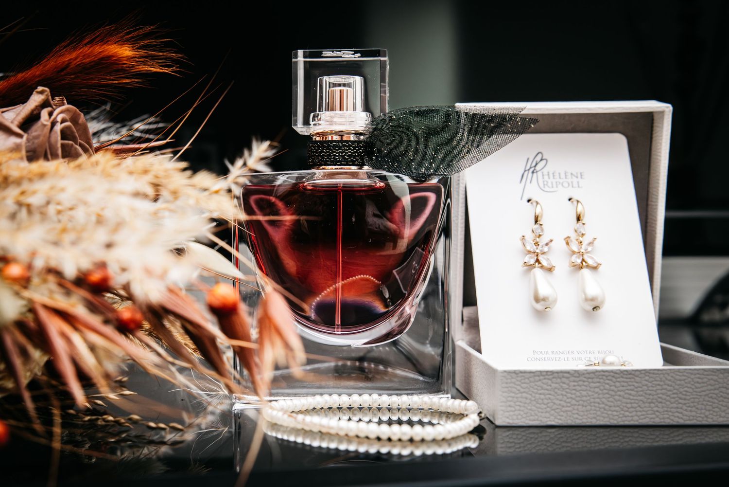 Flacon de parfum sombre élégant avec des boucles d’oreilles en perles dans une boîte‑cadeau et des fleurs séchées sur fond sombre.