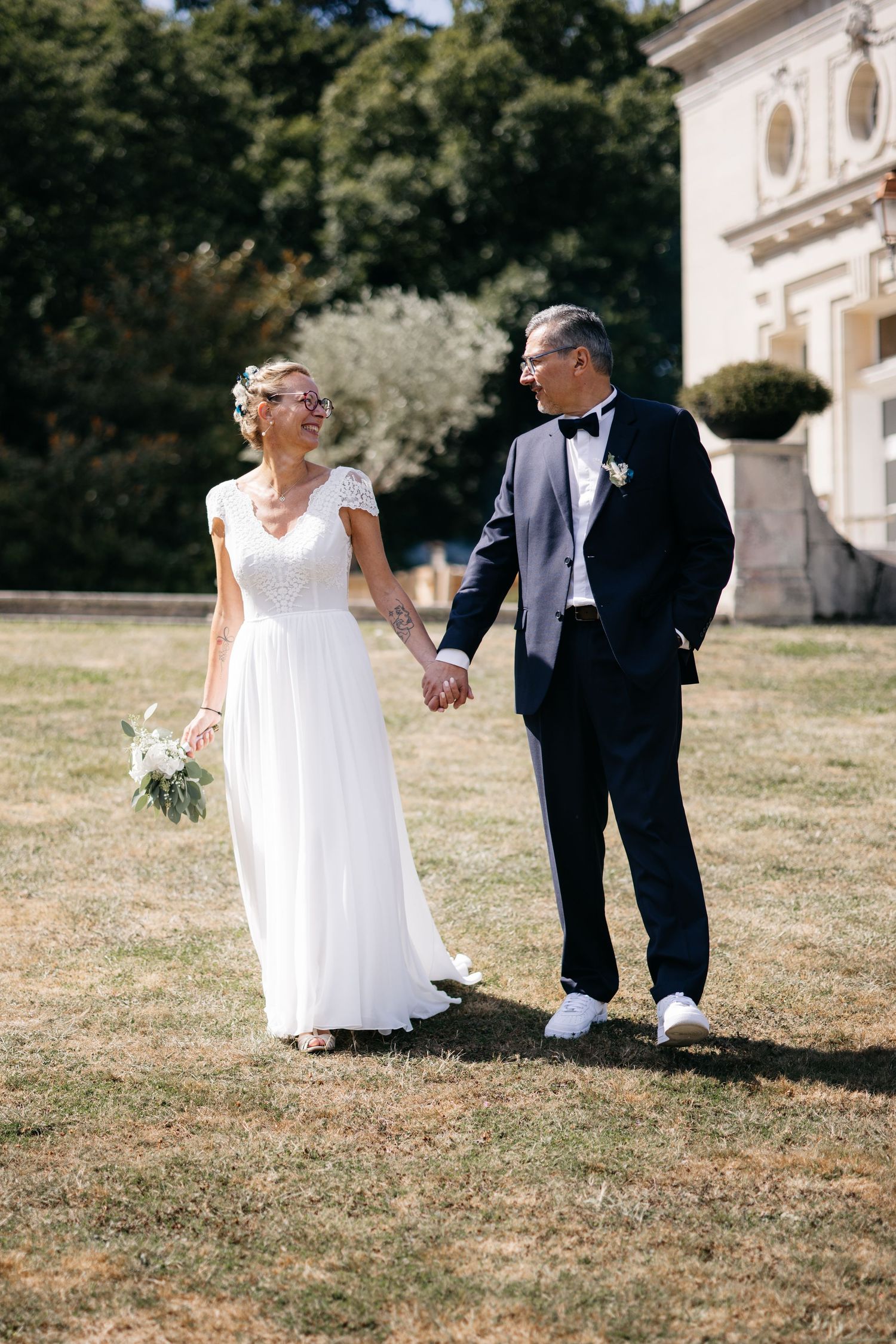 Une mariée dans une robe blanche fluide et un marié en smoking noir se tiennent la main sur la pelouse d'un domaine élégant.