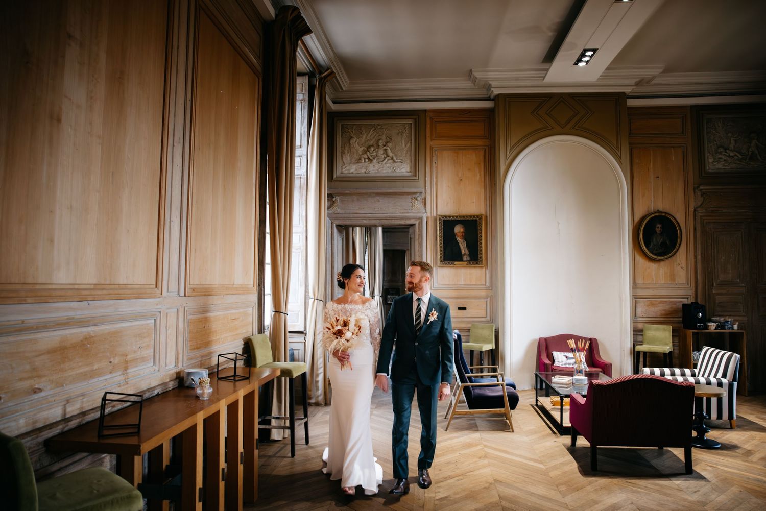 Un couple en tenue de mariage se tient dans une élégante pièce lambrissées avec des meubles vintage et une porte arquée.