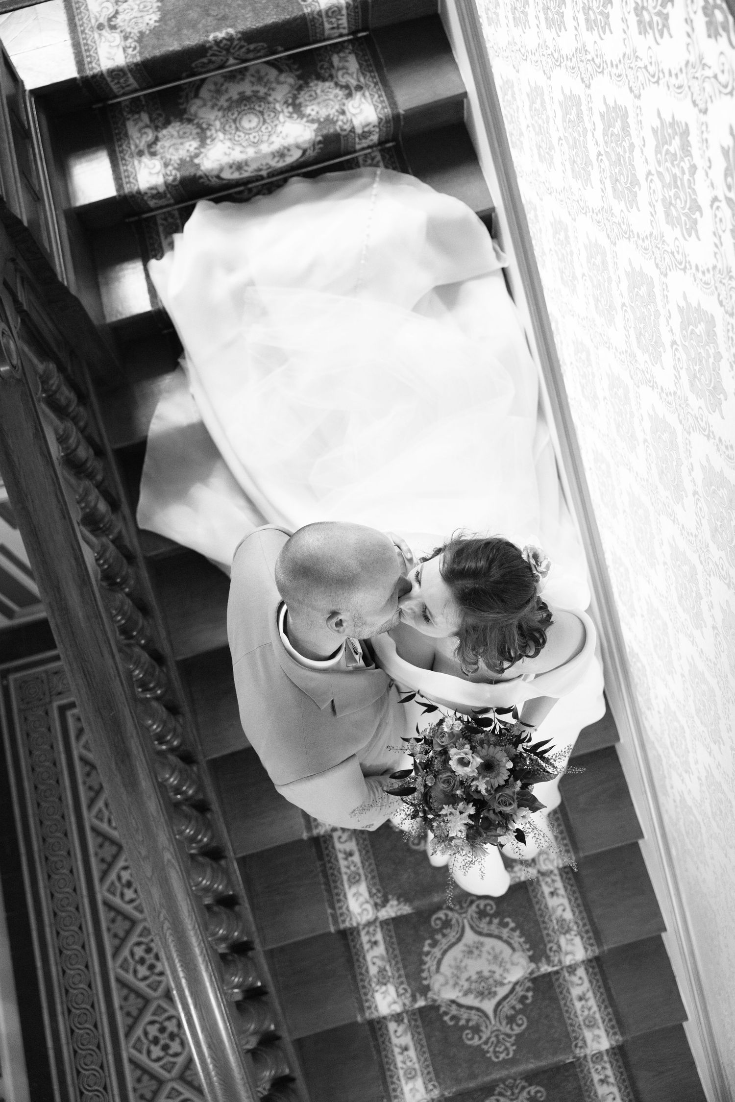 Vue aérienne en noir et blanc des mariés s'embrassant sur un escalier orné, la mariée tenant un bouquet de fleurs.