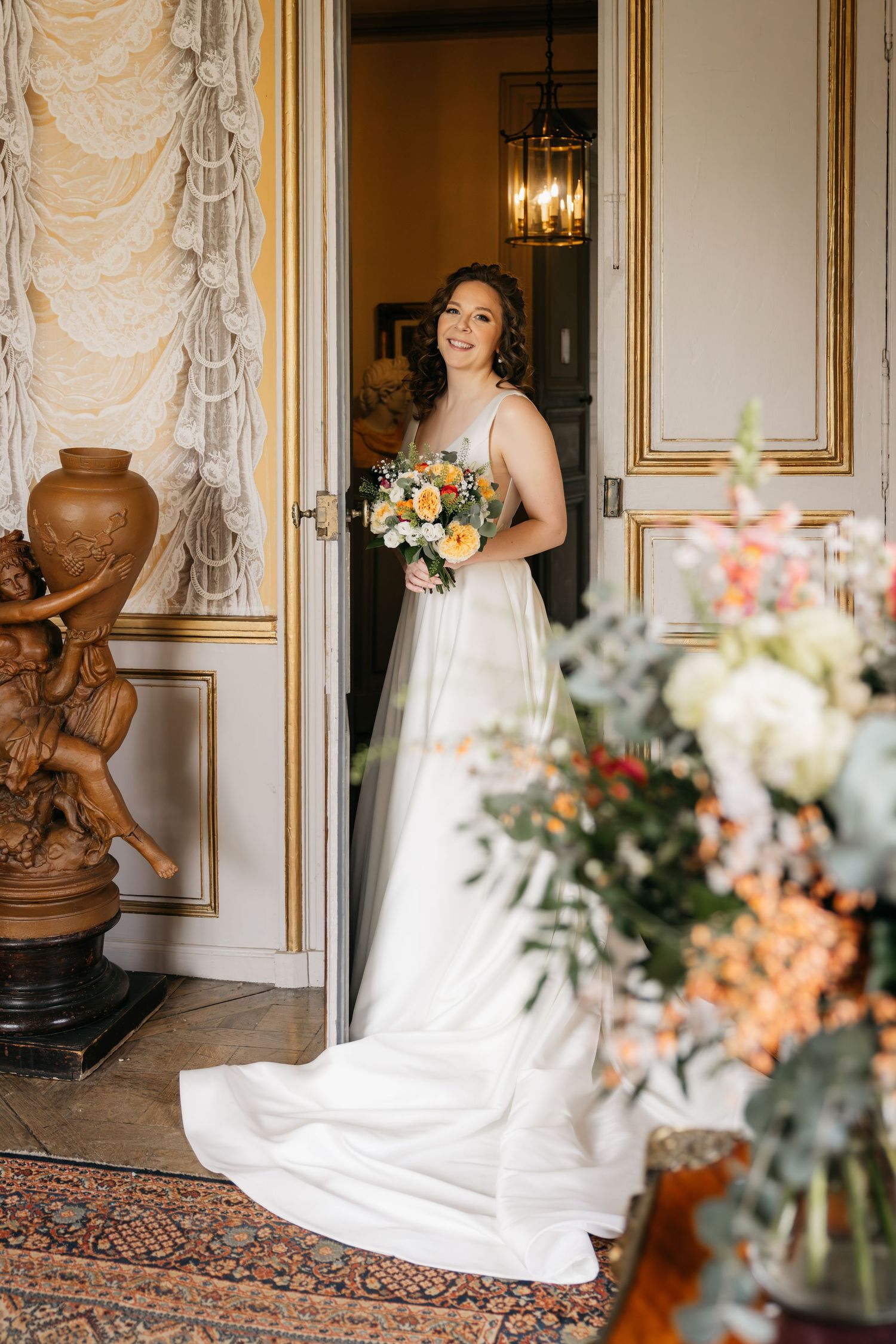 Mariée souriante en robe blanche à traîne tenant un bouquet coloré, debout dans une porte élégante aux bordures dorées