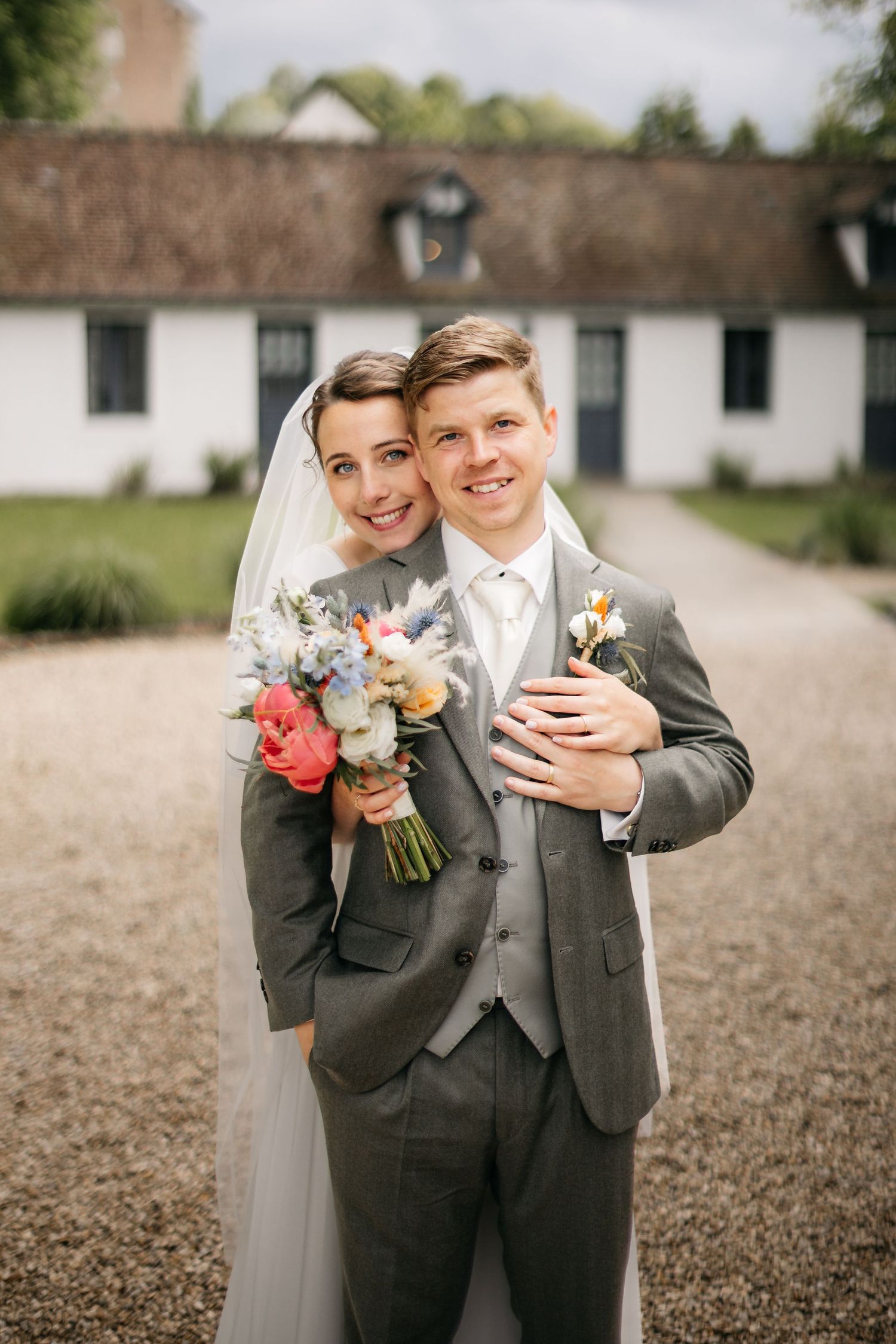Mariés souriants en extérieur, la mariée avec un bouquet coloré, le marié en costume gris, bâtiment blanc derrière.