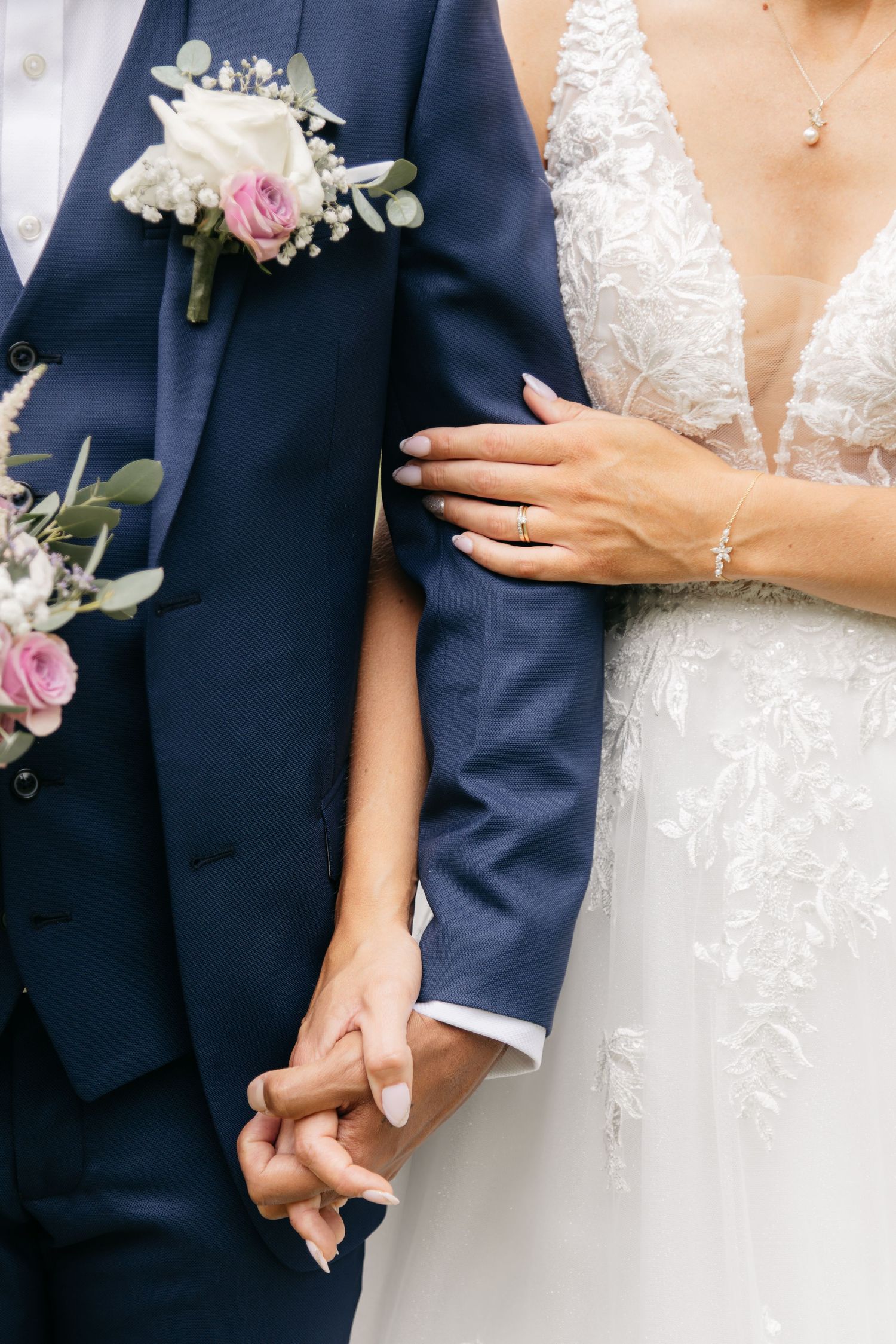 Mariés se tenant par la main, la mariée portant une robe de mariée en dentelle avec une bague en diamant, le marié vêtu d’un costume bleu