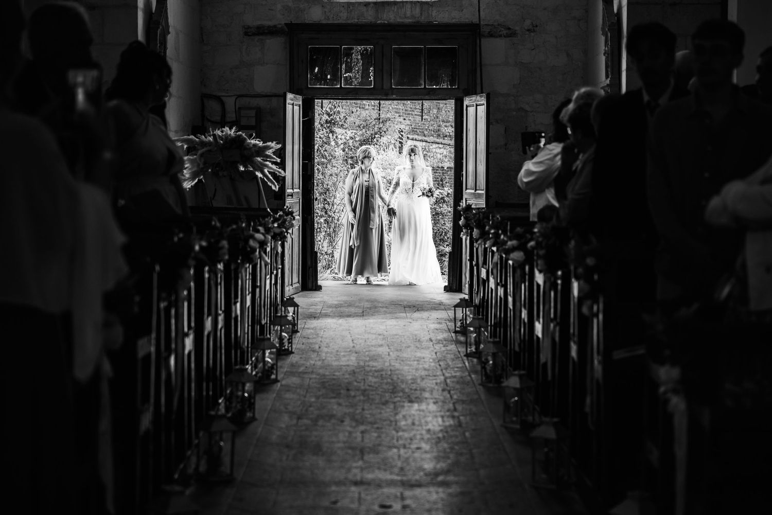 Photo de mariage en noir et blanc montrant la mariée et le marié debout devant les portes d’entrée de l’église, avec des invités assis.