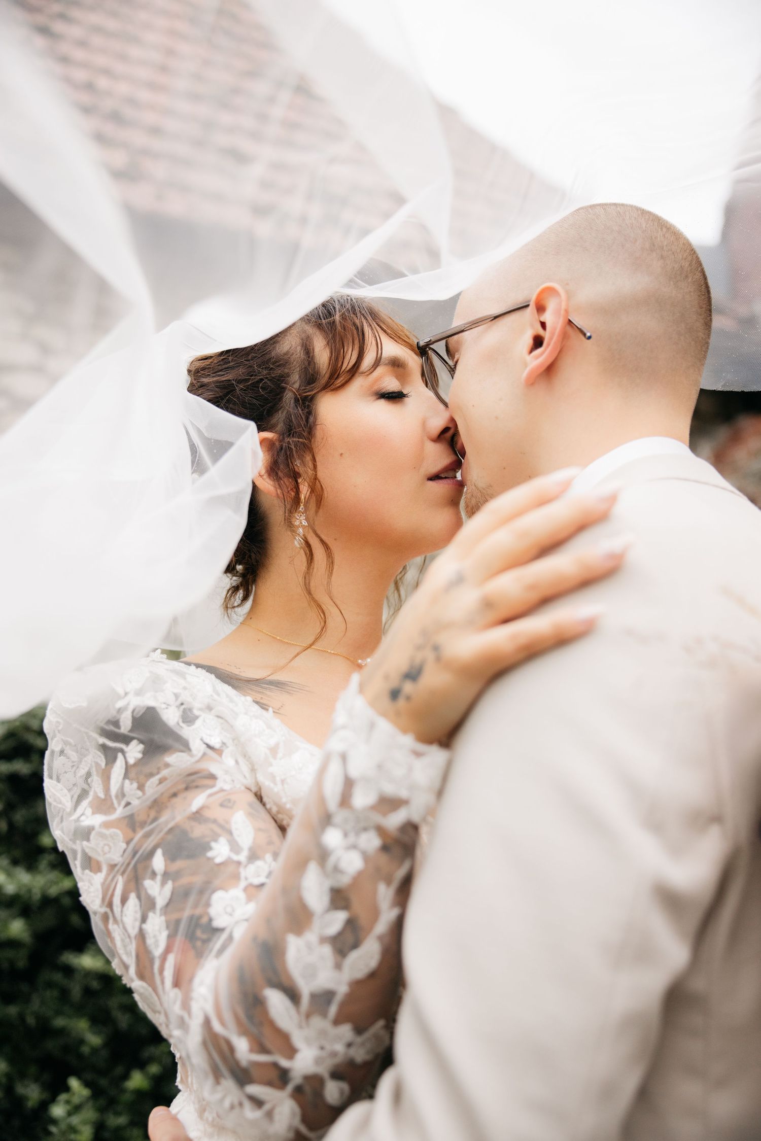 Les mariés partageant un baiser romantique sous un voile blanc fluide, la mariée portant une robe de mariée en dentelle.