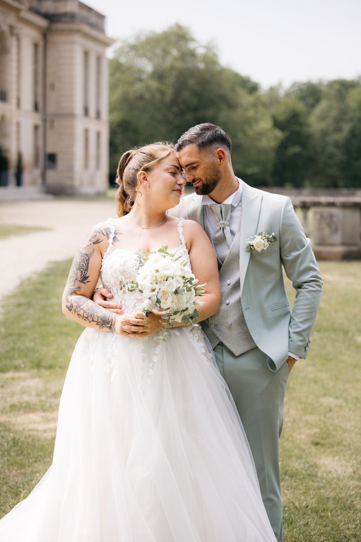 Les mariés s'enlacent à l'extérieur, près d'un bâtiment élégant ; la mariée tient un bouquet blanc dans sa robe de dentelle