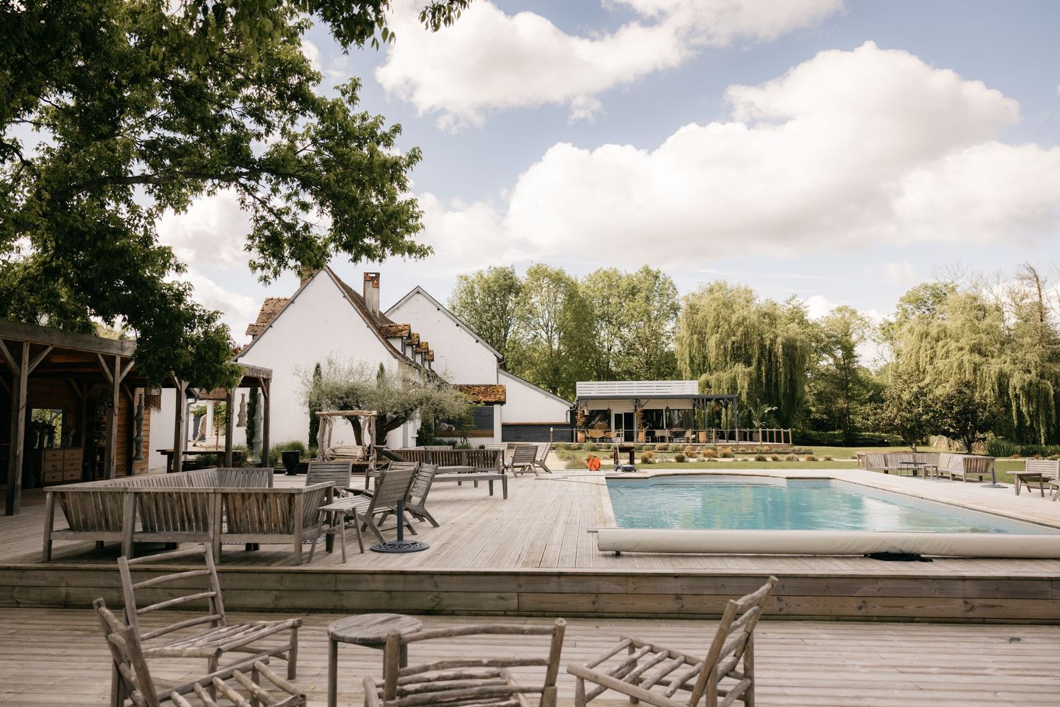 Élégante piscine extérieure avec terrasse en bois, mobilier de patio et arbres luxuriants dans un domaine champêtre.