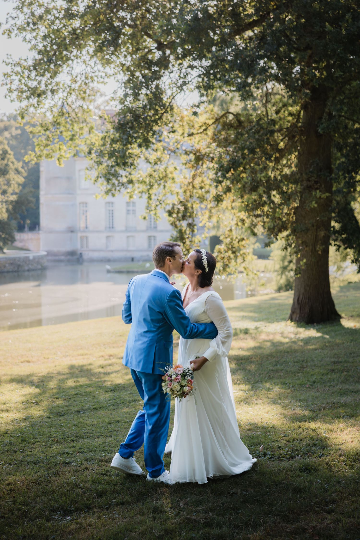 La mariée en robe blanche fluide et le marié en costume bleu s'embrassent sur une pelouse ensoleillée avec un élégant manoir en arrière-plan