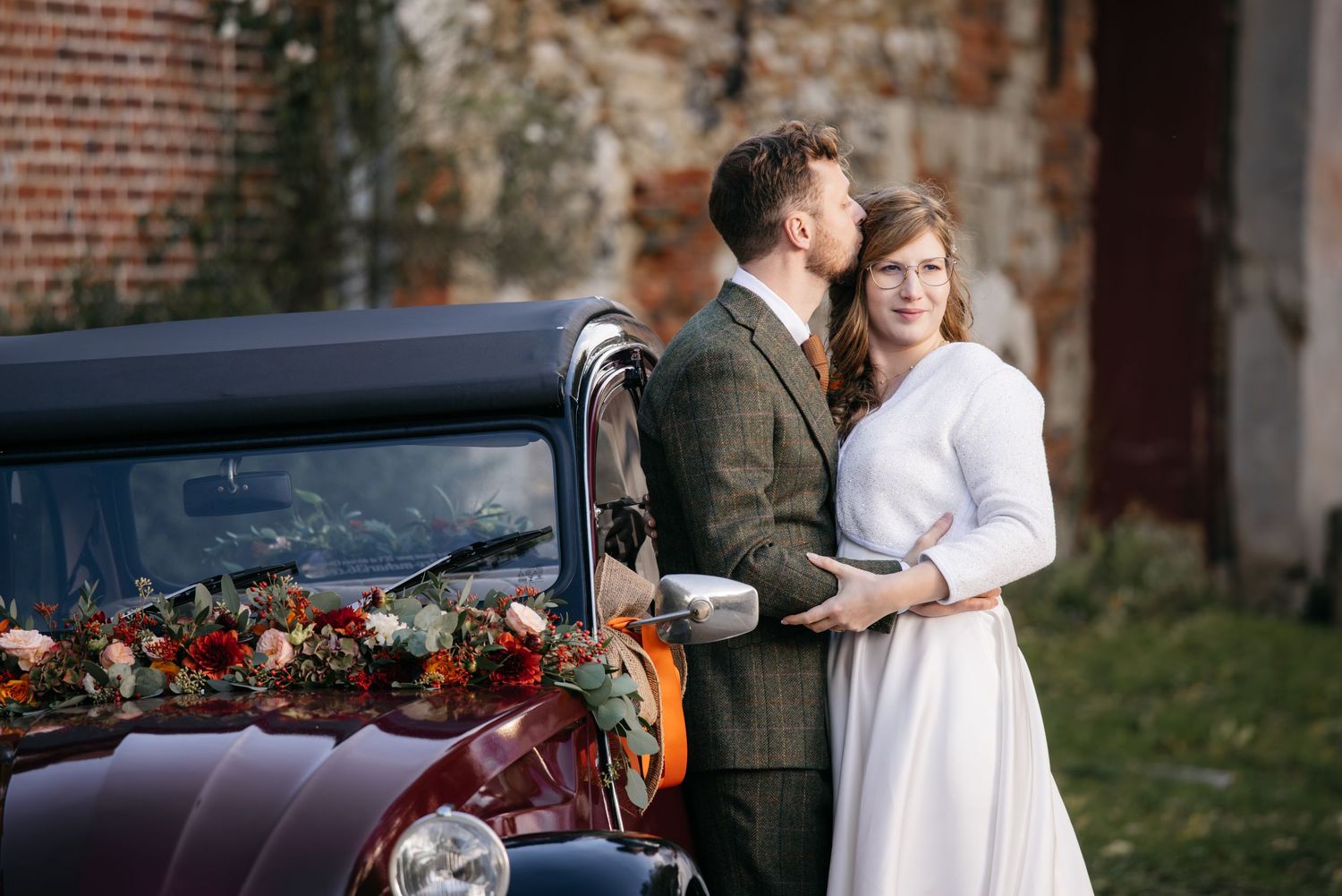 Un couple en tenue de mariage pose pour des photos romantiques devant des murs de briques rustiques et des voitures anciennes.