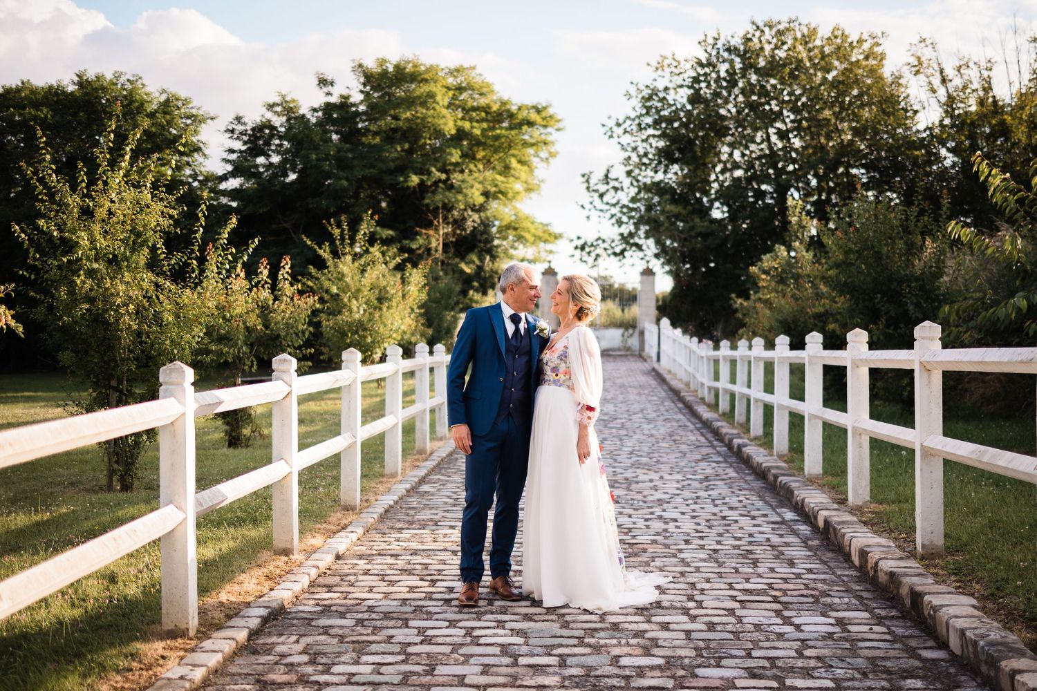 A bride and groom standing together on a romantic cobblestone path lined with white fence posts at sunset.