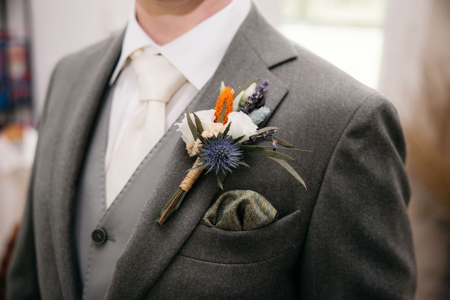 Gros plan sur le costume gris d'un marié avec une boutonnière de chardon bleu, fleurs séchées et pochette en tweed.