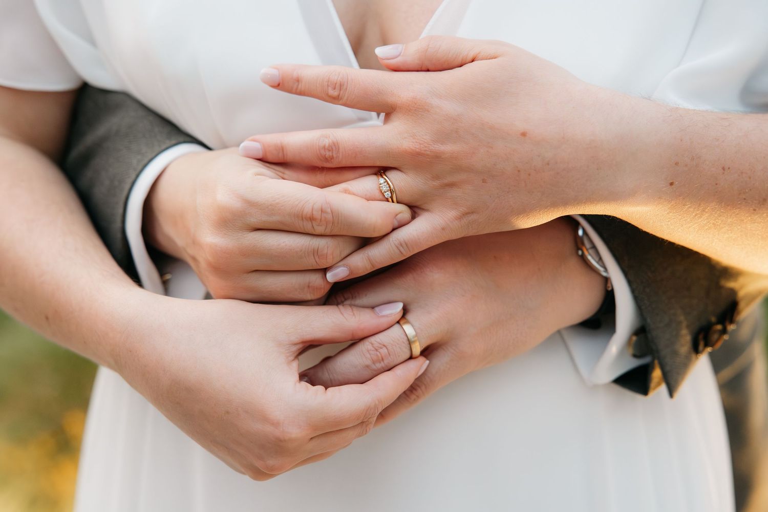 Gros plan sur les jeunes mariés se tenant par la main montrant leurs alliances dorées, la mariée en robe blanche, le marié en costume gris.
