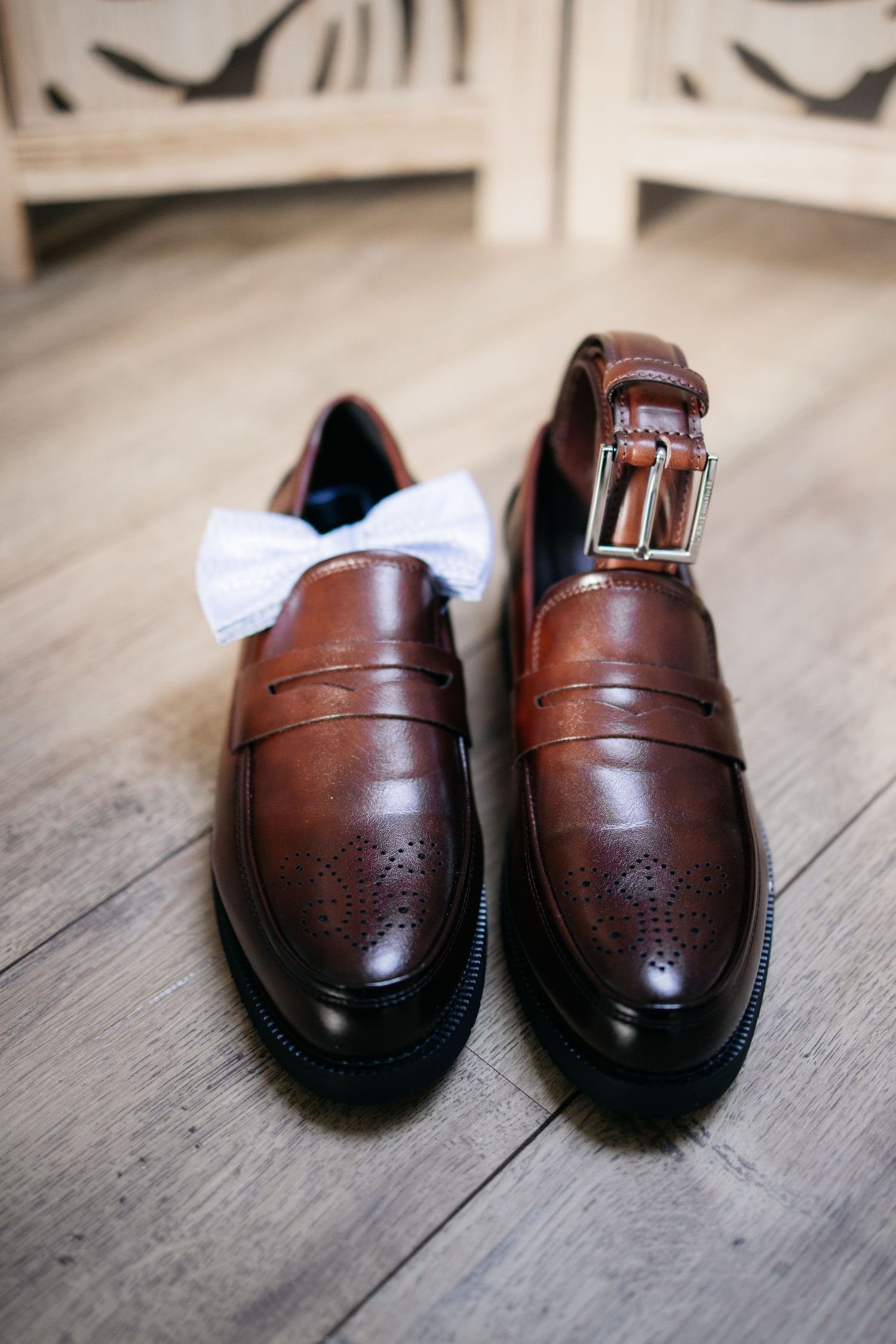Mocassins en cuir marron penny loafers avec un nœud papillon blanc et une ceinture dorée posés sur un sol en bois.