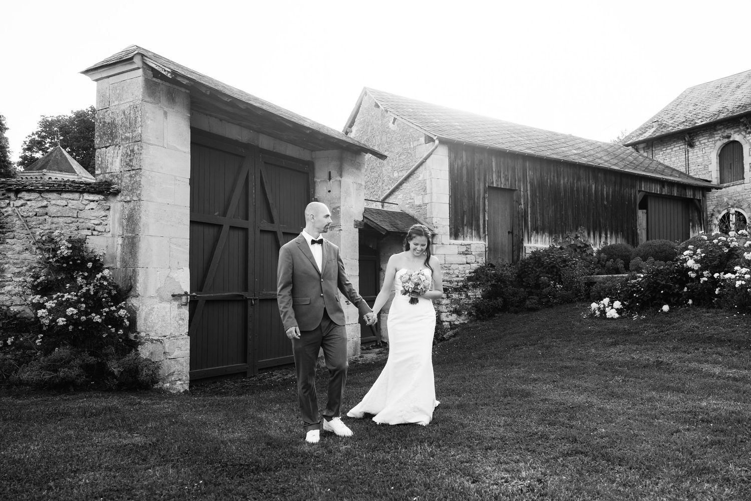 Photo en noir et blanc des mariés se tenant par la main devant une grange rustique en pierre le jour de leur mariage.