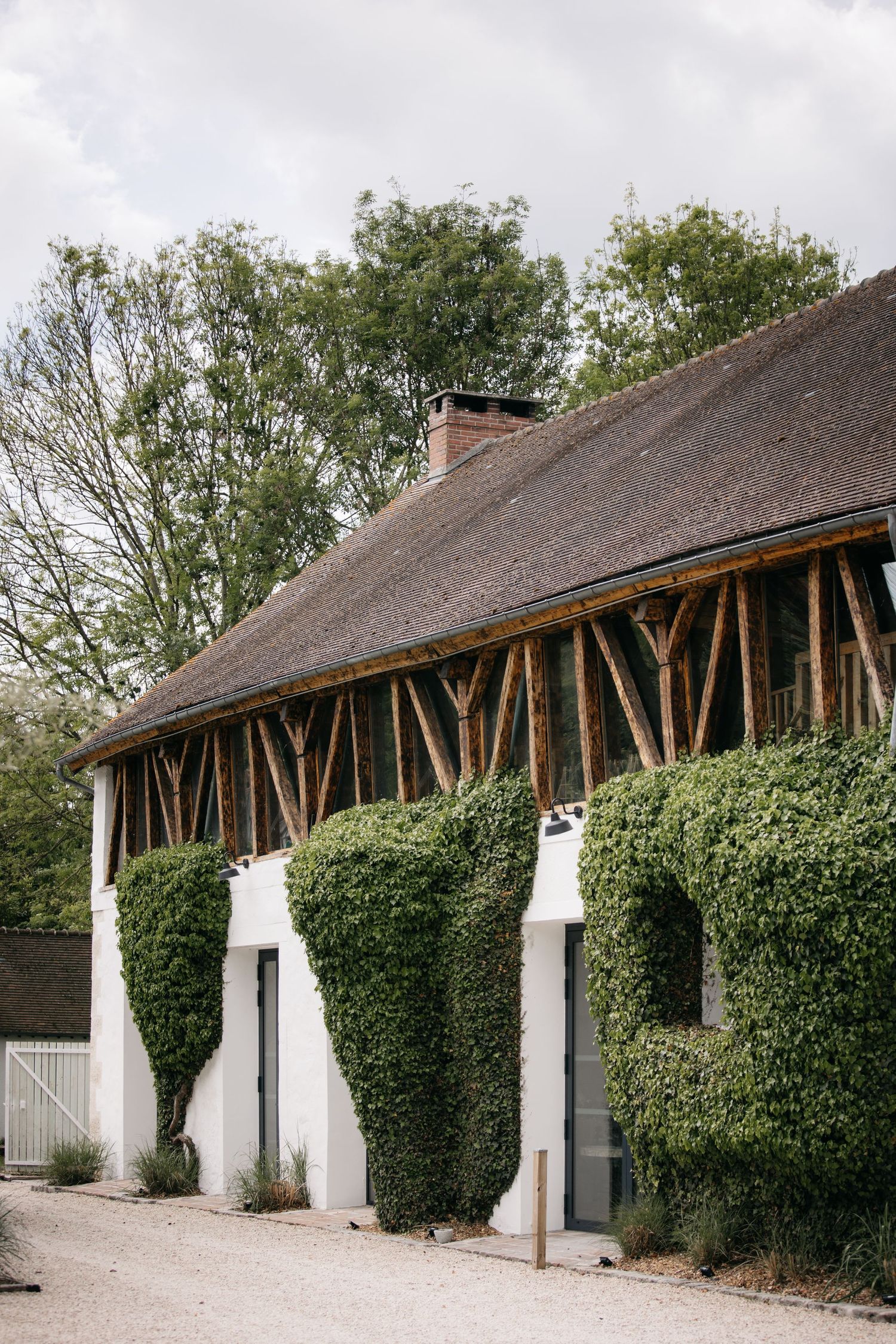 Bâtiment rustique à colombages, murs blancs couverts de lierre, toit de chaume et cheminée, entouré d'arbres verdoyants.