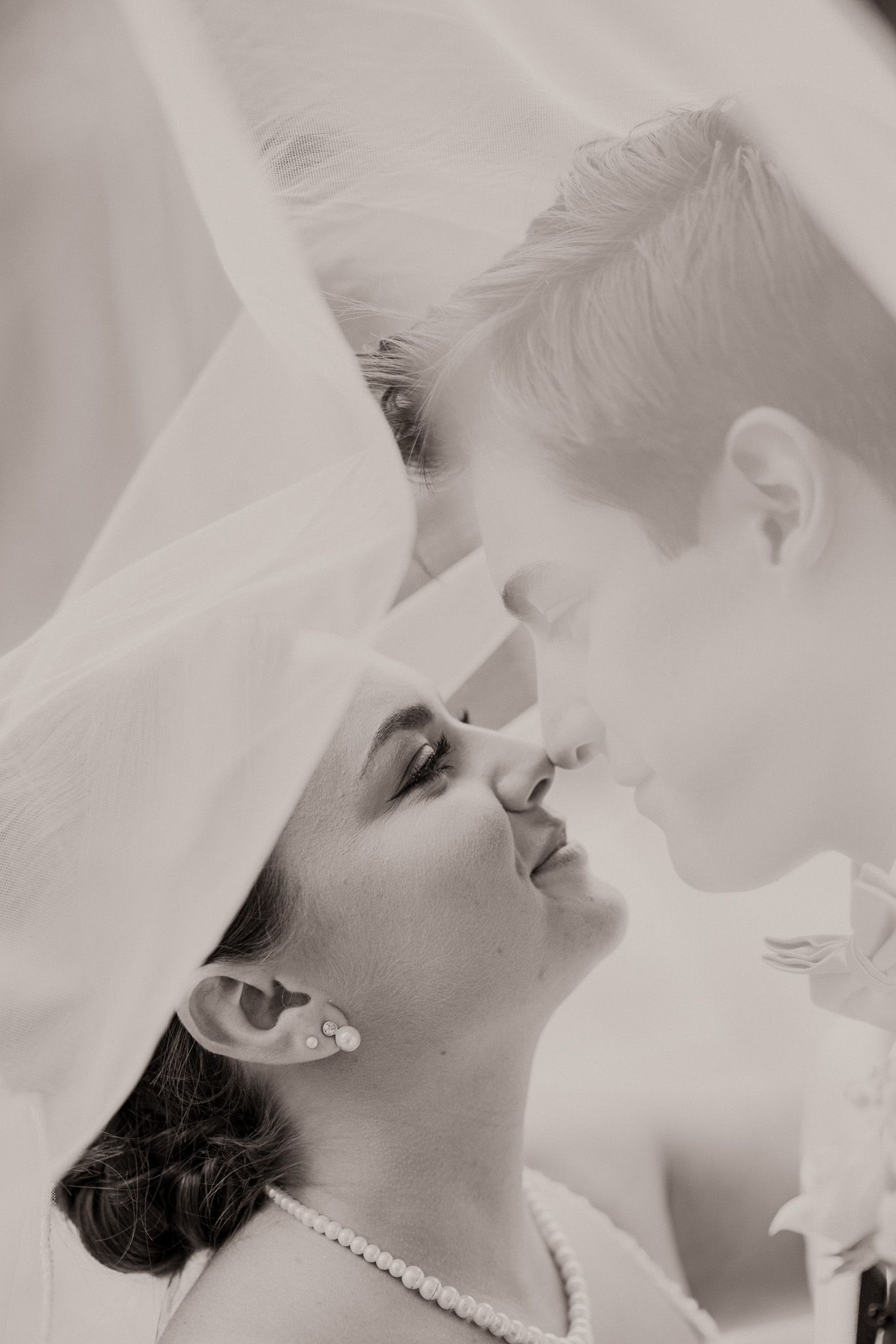 Black and white wedding photo of two brides sharing a tender moment under a veil, wearing pearls.