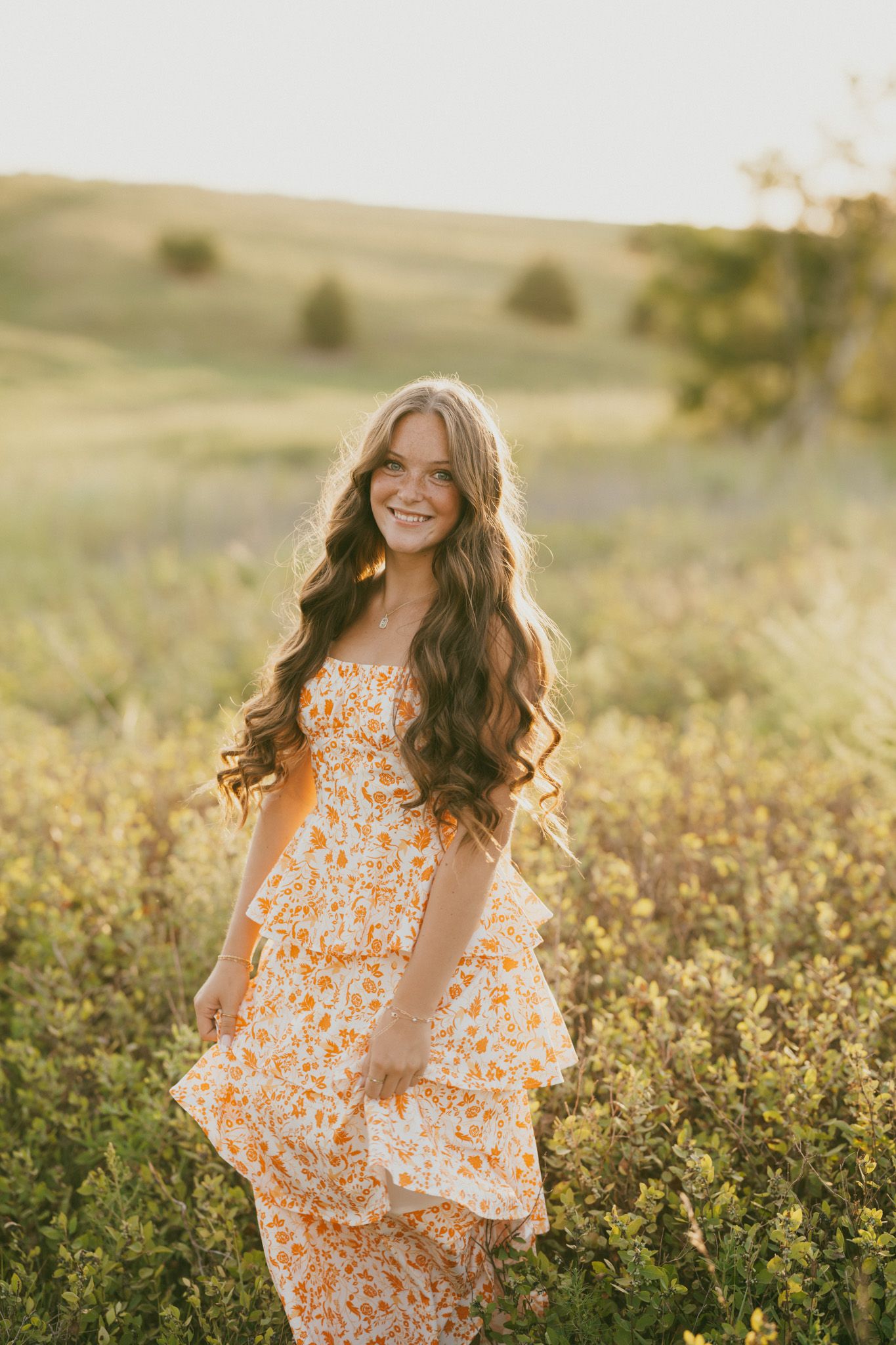 Young woman in floral orange sundress smiling in golden hour sunlight among wildflowers in a green meadow.