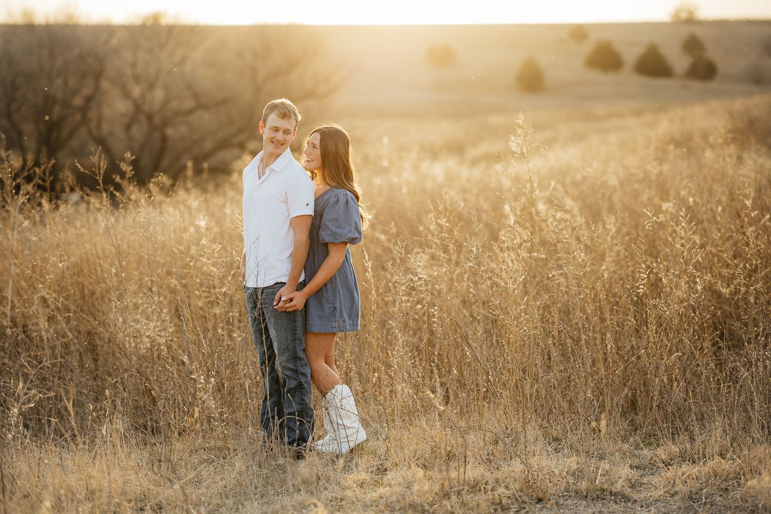 Couple holding hands in golden field at sunset, man in white shirt and woman in blue dress with white boots.