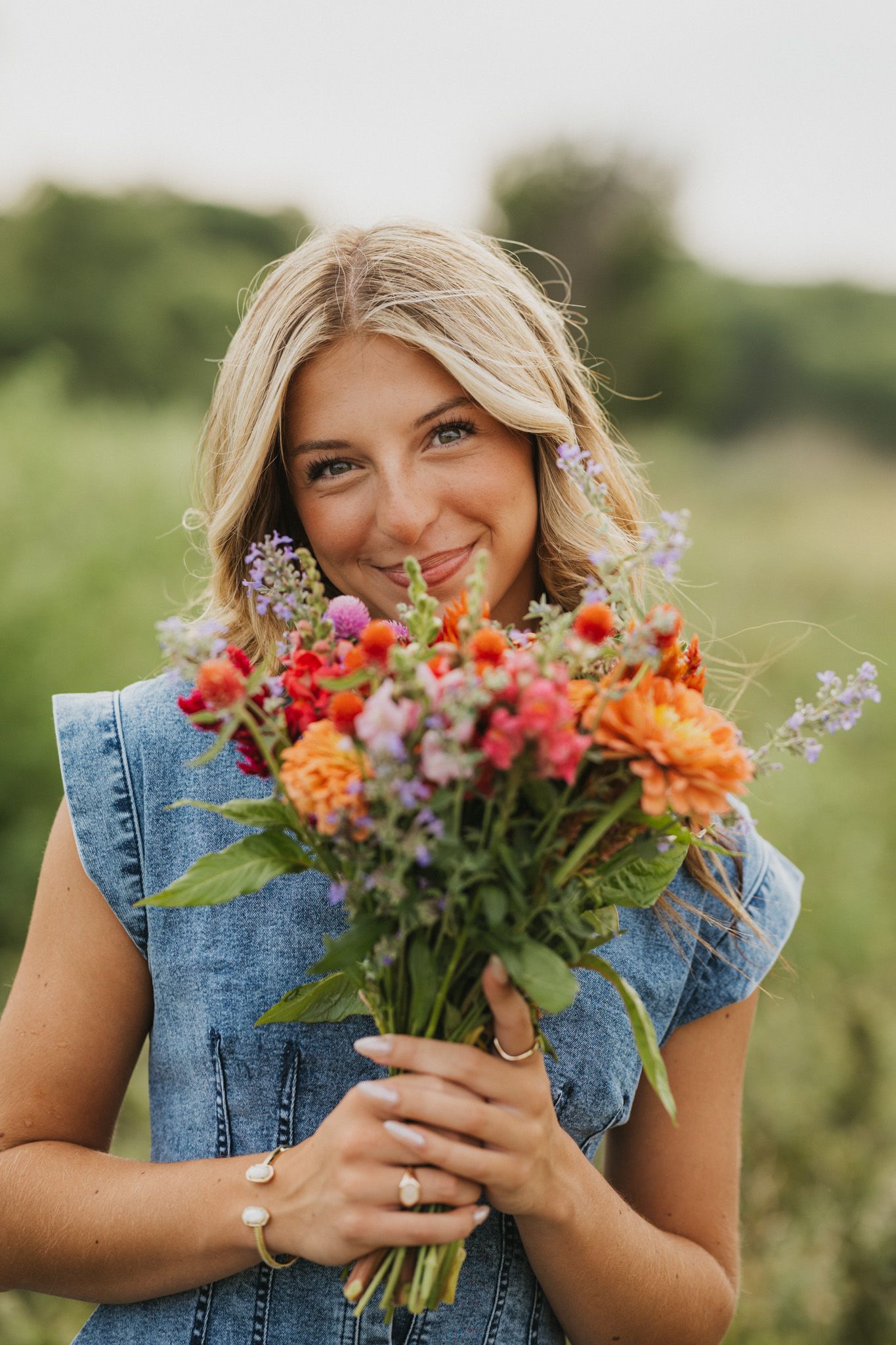 Smiling blonde woman in denim vest holding colorful wildflower bouquet outdoors in green field.
