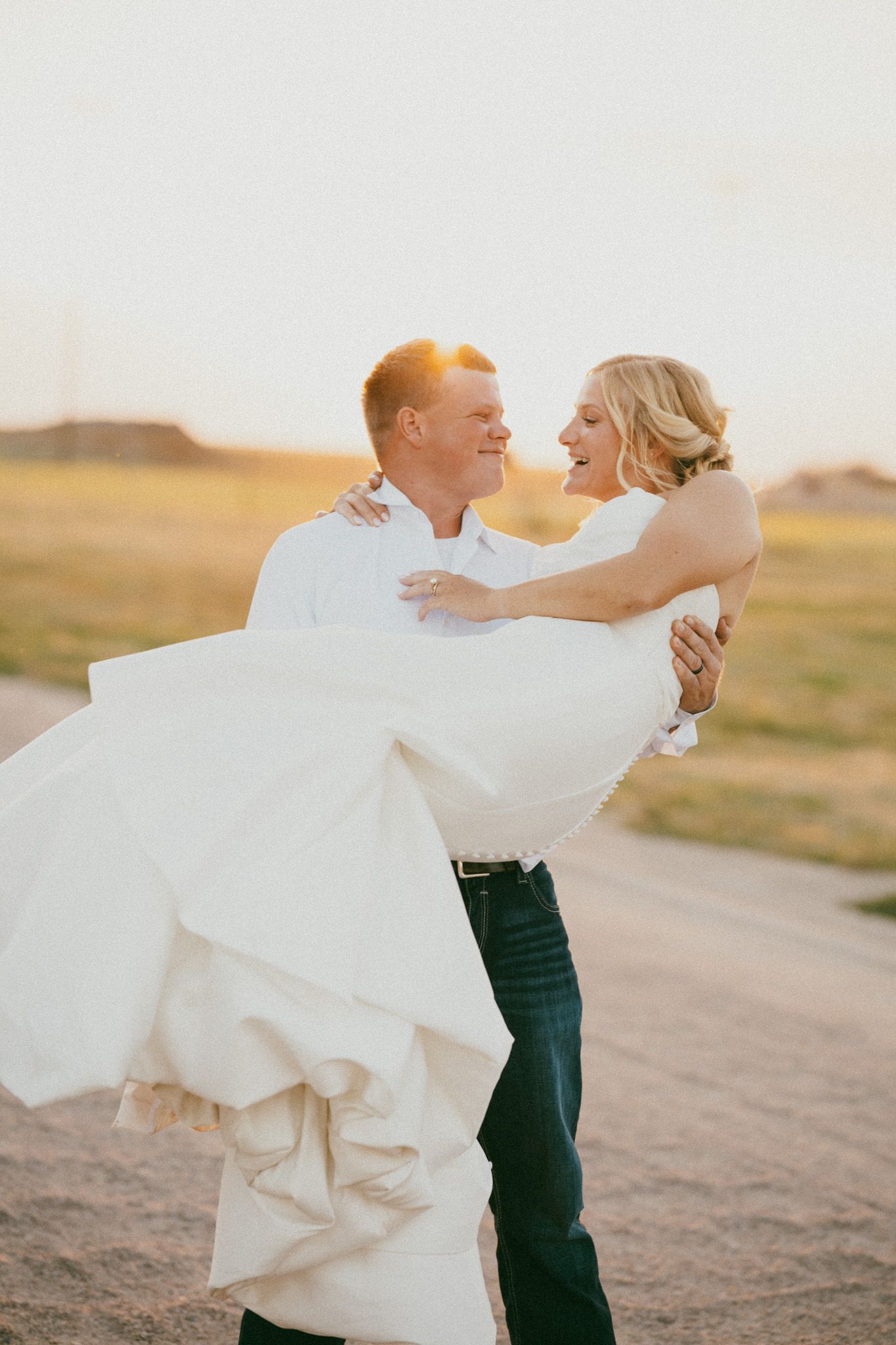 Groom in white shirt and jeans lifts smiling bride in white wedding dress during golden hour sunset outdoors.