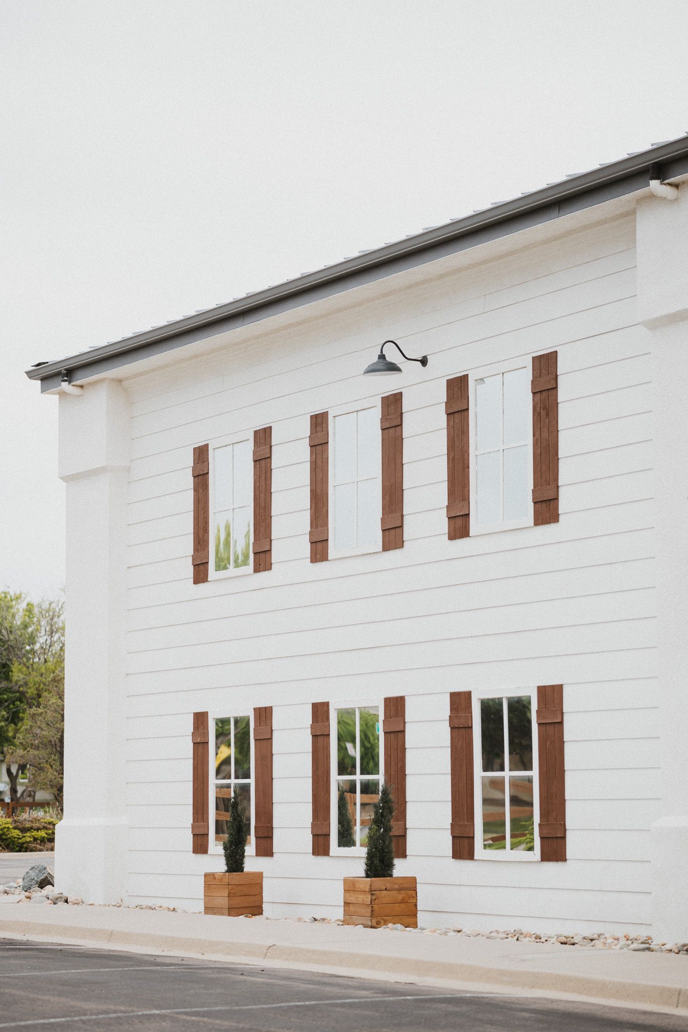 White farmhouse building with brown wood shutters, black gooseneck lights, and potted topiaries flanking lower windows.