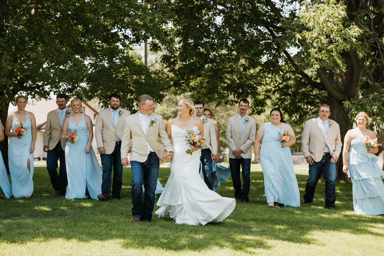 Wedding party walks outdoors in jeans, bridesmaids in light blue gowns, groomsmen in tan suits, bride in white dress.