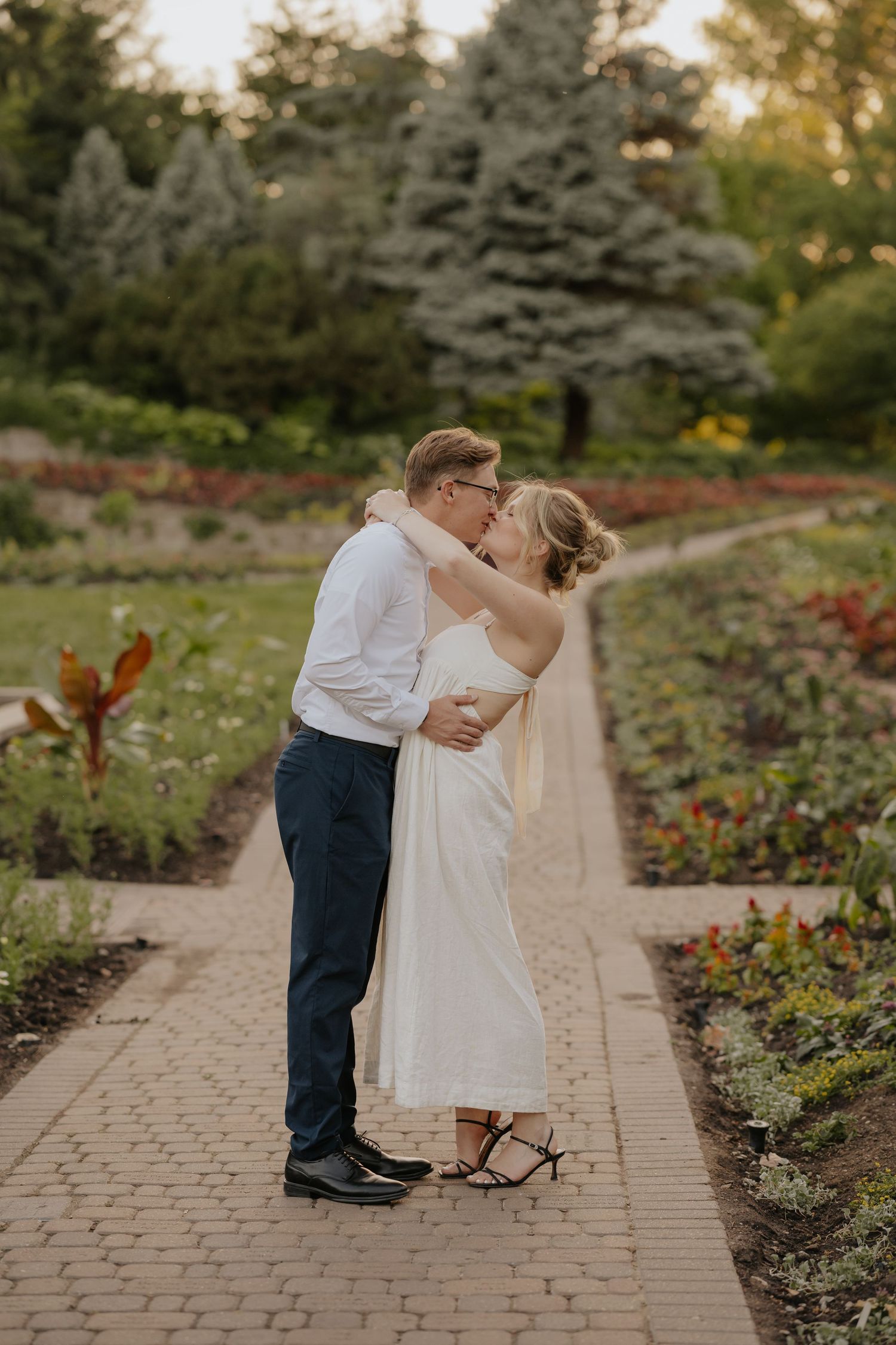 A couple shares a romantic kiss on a brick garden path surrounded by colorful flowers and lush greenery.