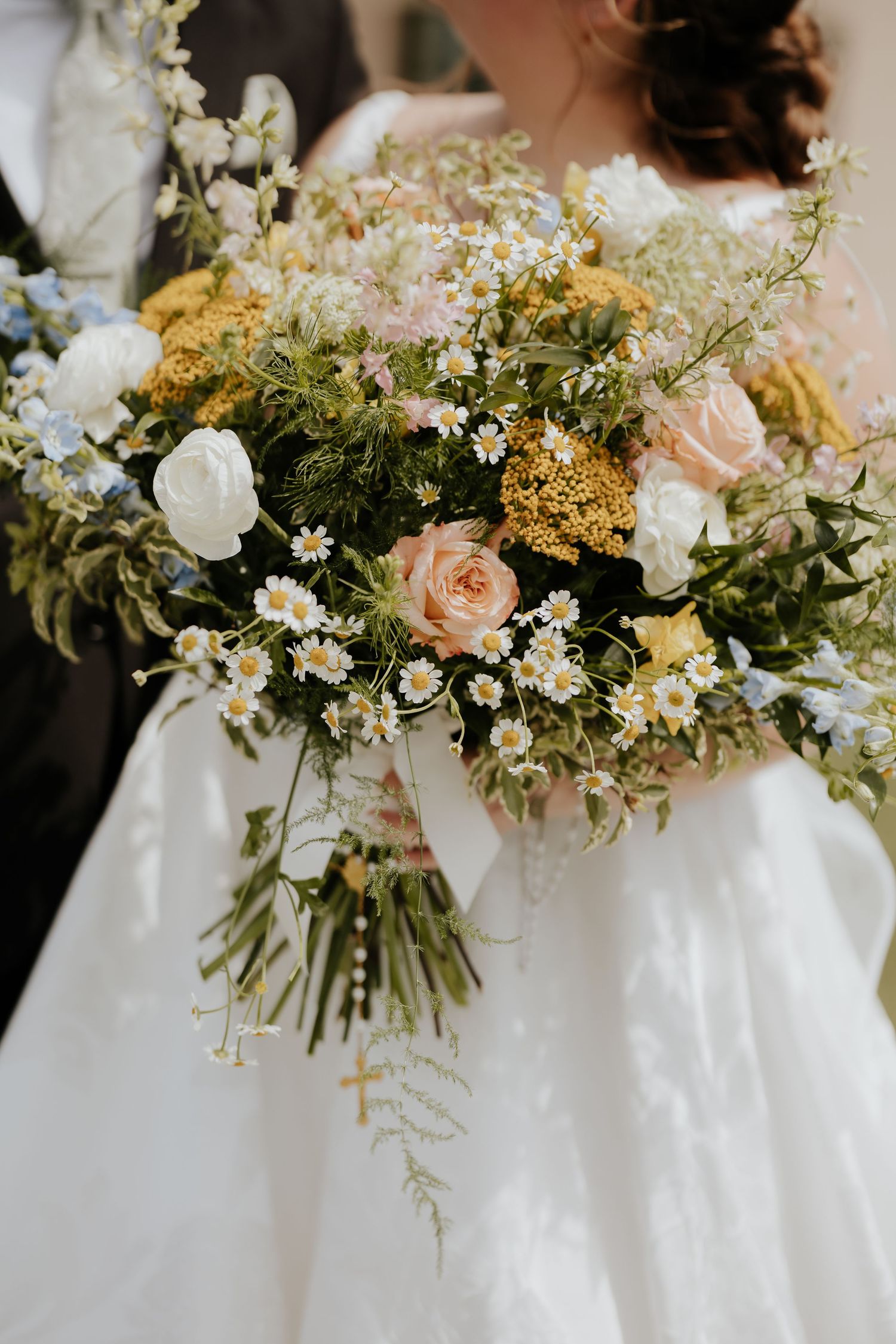 Bride holding a lush wildflower wedding bouquet with roses, daisies, and yellow blooms against a white gown.