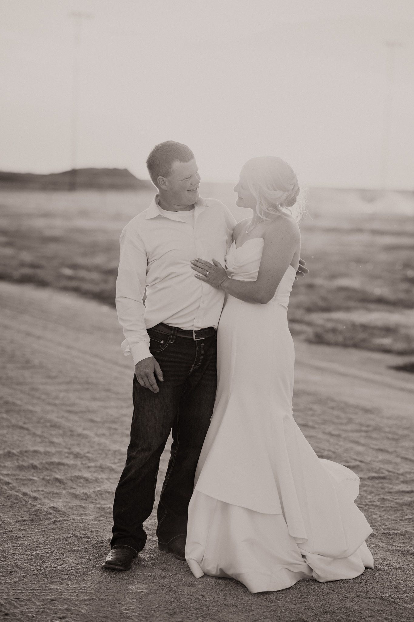 Black and white photo of a bride in a white gown and groom in jeans embracing on a sandy beach at sunset.