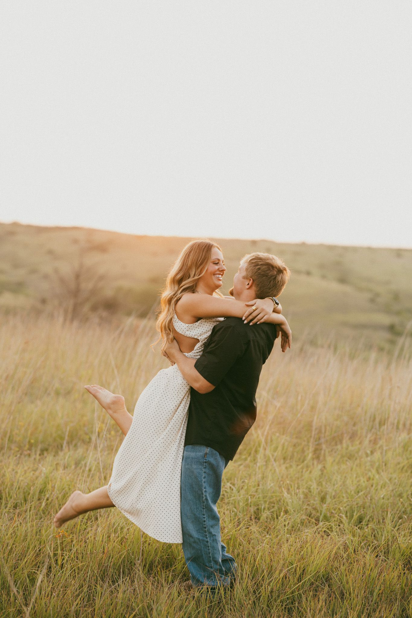 A joyful couple embraces in a sunlit field, the man lifting the smiling woman in a white dress at golden hour.