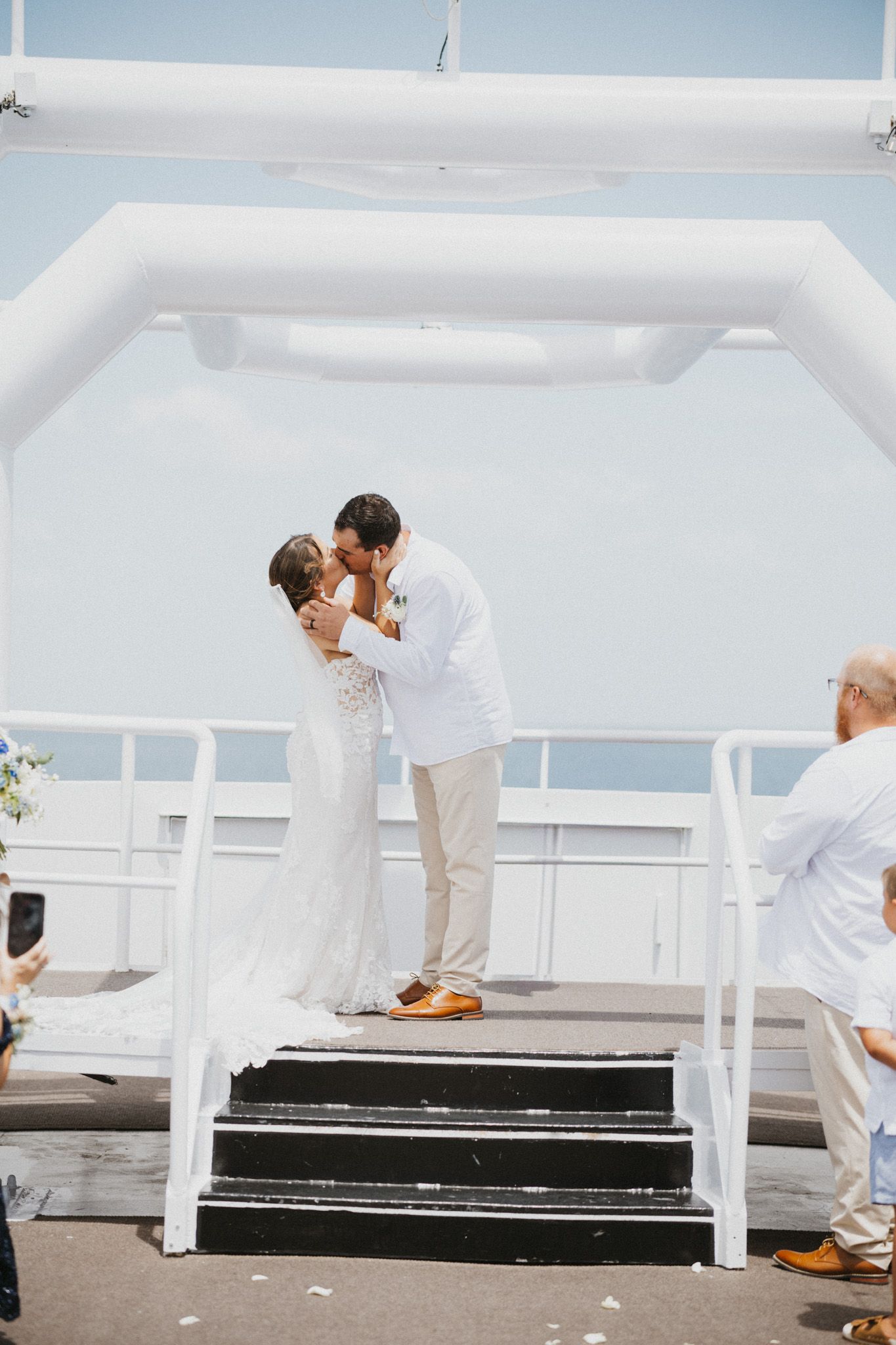 Bride and groom share first kiss on ship deck during outdoor wedding ceremony with ocean backdrop.