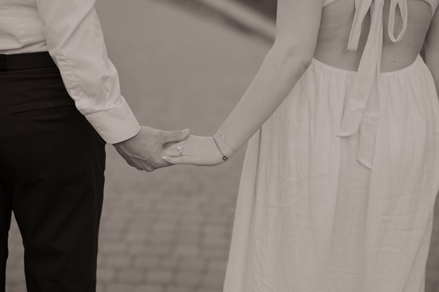 Black and white photo of a couple holding hands, woman in white dress, romantic outdoor setting.