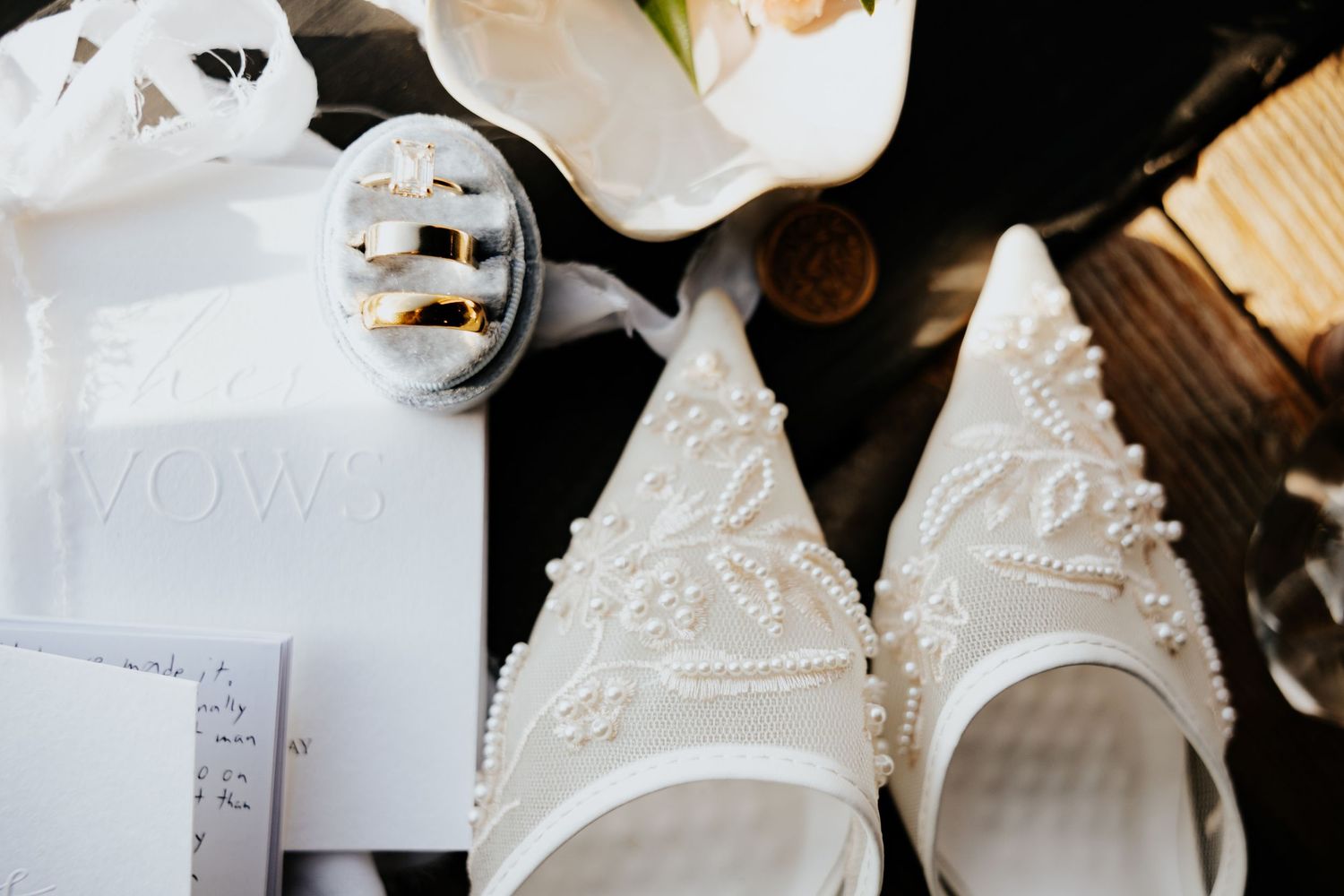 Elegant wedding flat lay featuring pearl-embellished bridal shoes, gold rings, vow books, and white florals on dark wood.