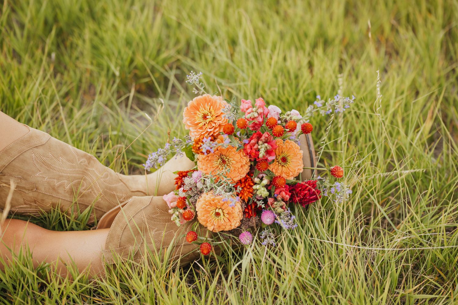 Hands holding a vibrant bouquet of orange dahlias, red and pink flowers in lush green grass.