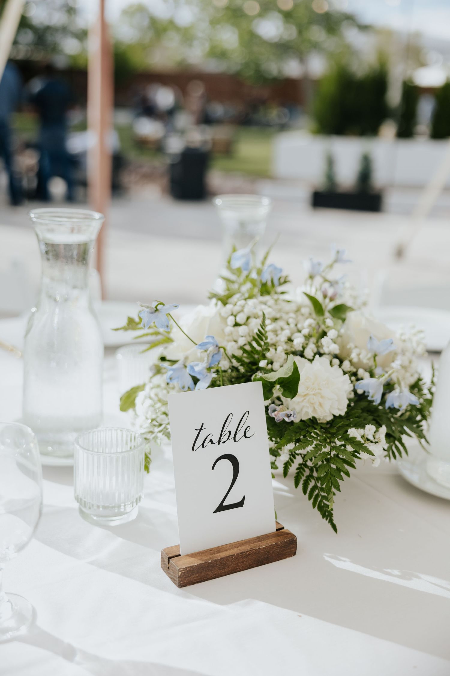 Elegant wedding table 2 centerpiece with white and blue flowers, fern greenery, glass carafe, and ribbed drinking glasses on white linen.