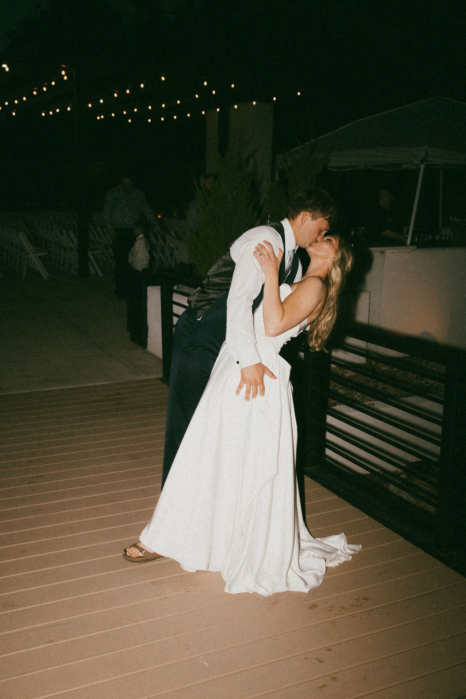 Bride and groom sharing a romantic dip kiss on an outdoor deck at night with string lights glowing above.