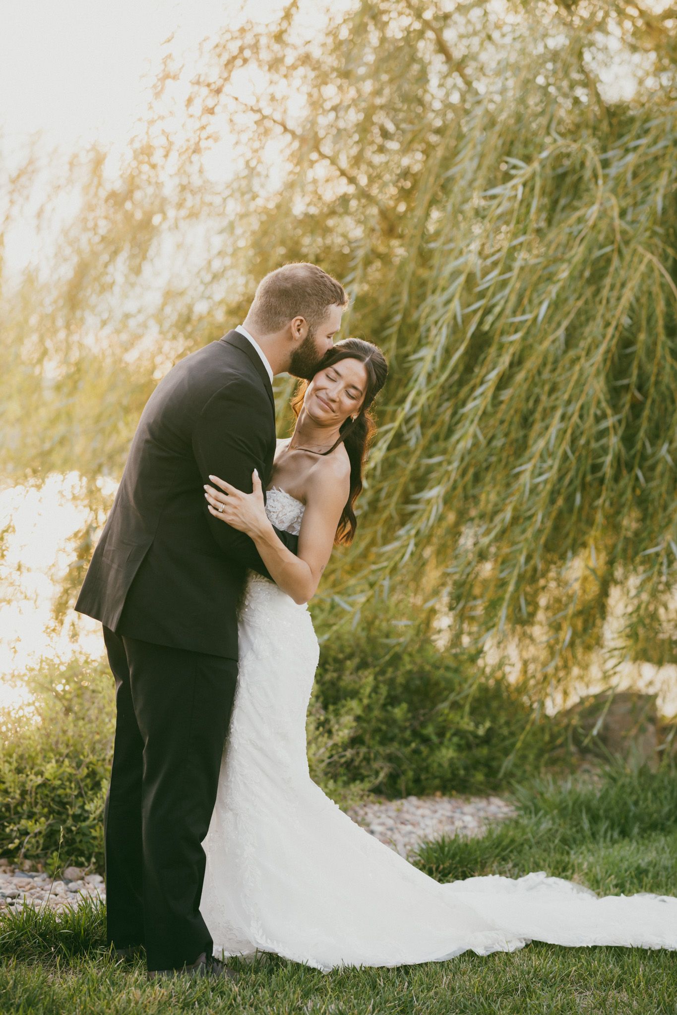 A groom in a dark suit kisses his bride in a white gown under a willow tree by a sunlit lake.